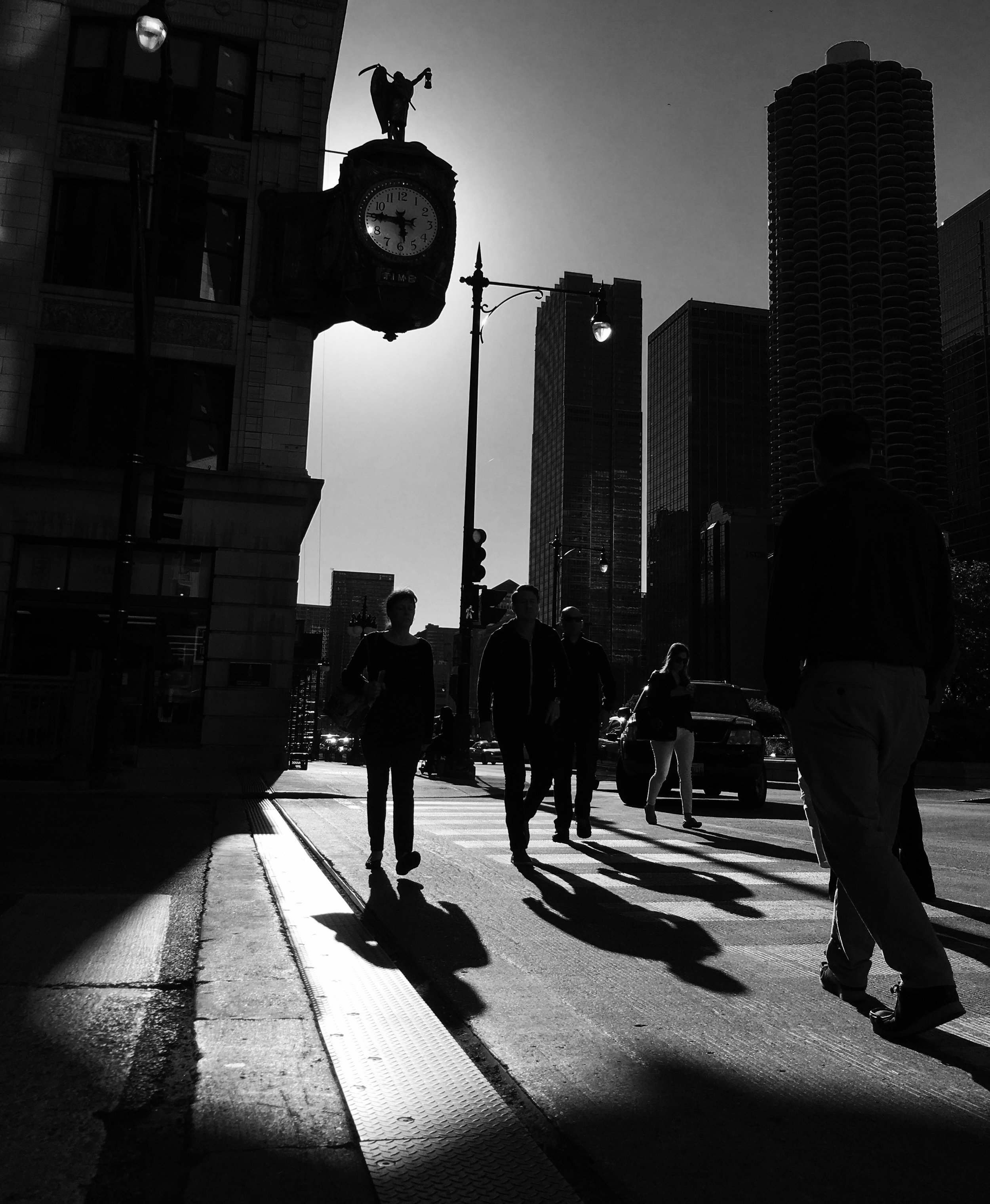 Silhouettes of people walking on a city sidewalk with a clock tower and high-rise buildings in the background, and streetlights illuminating the scene.