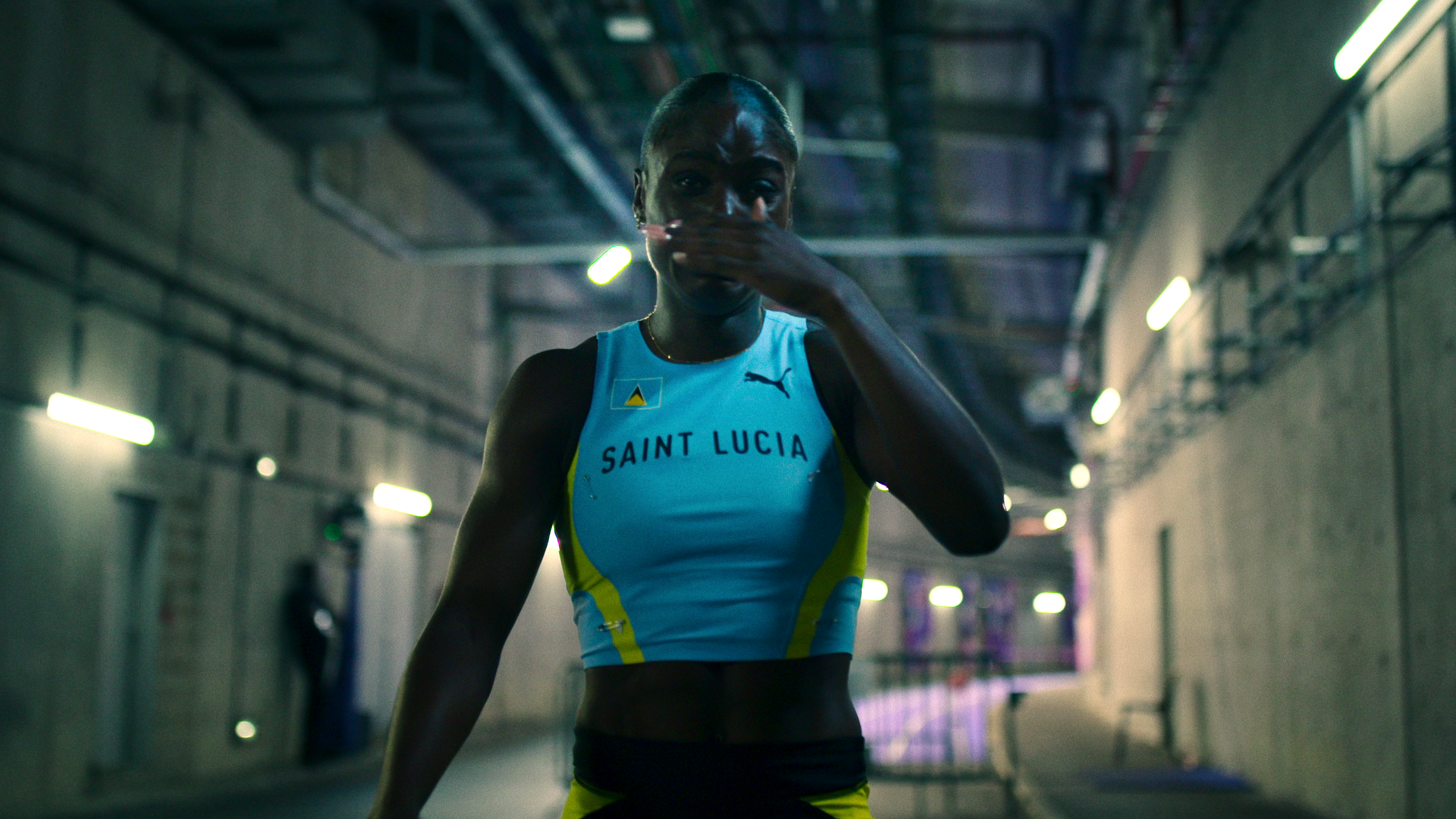 An athlete in Saint Lucia athletic gear places her hand over her mouth in a tunnel with industrial lighting.