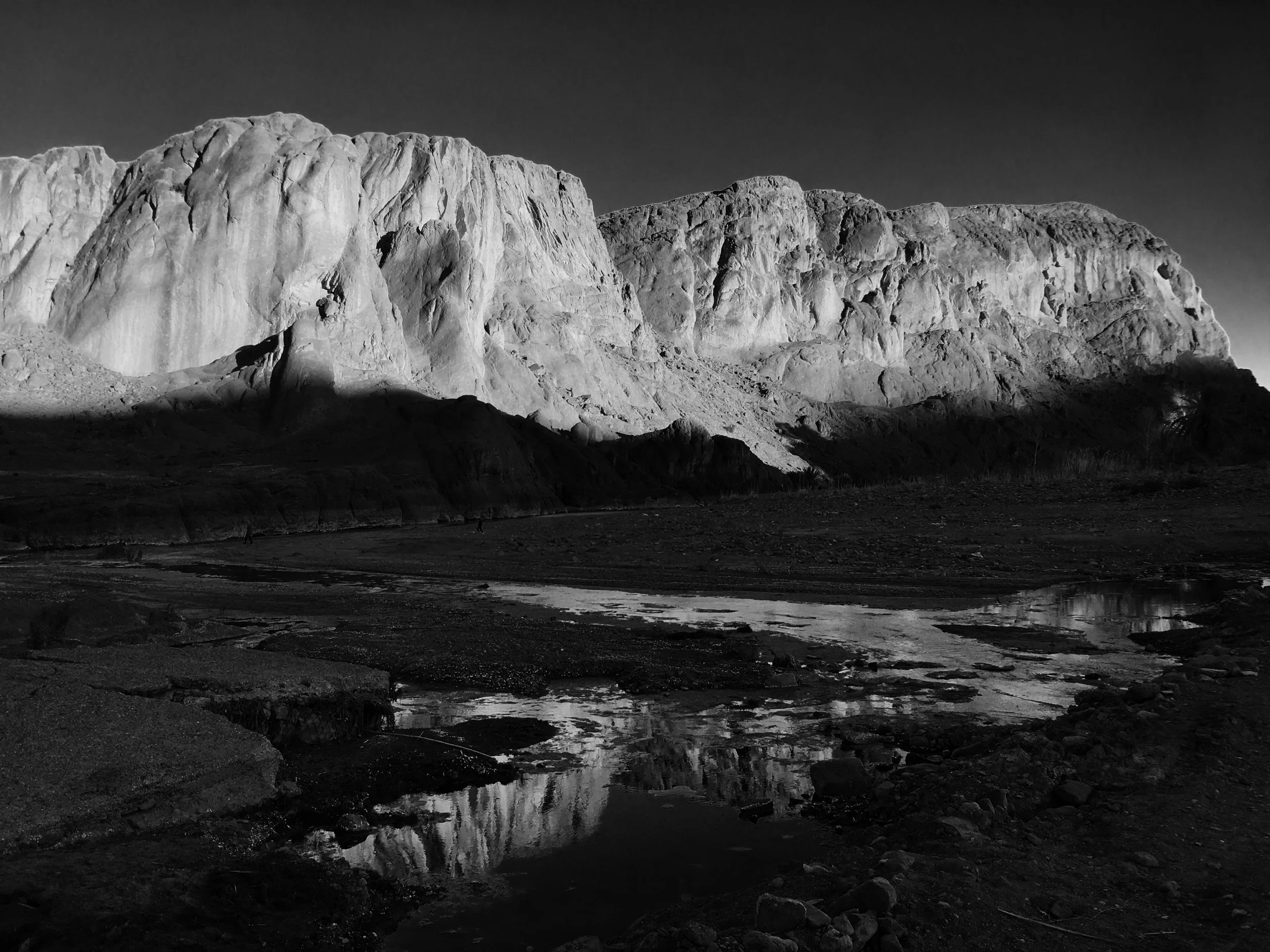 Black and white photo of large, rugged ice cliffs with a smooth water reflection at the base, under a dark sky.