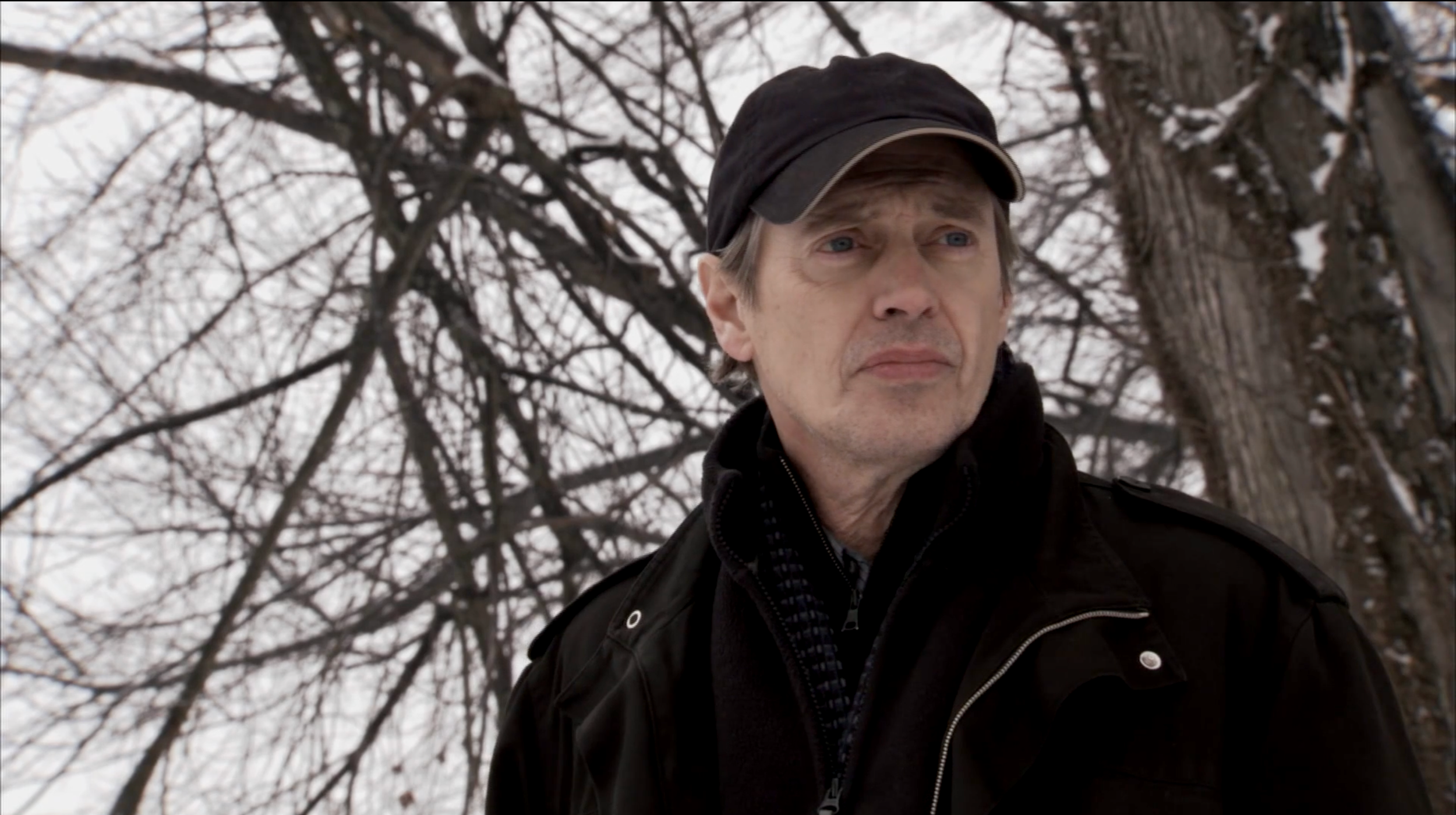 A man wearing a black cap and dark jacket standing outdoors on a winter day with leafless trees in the background