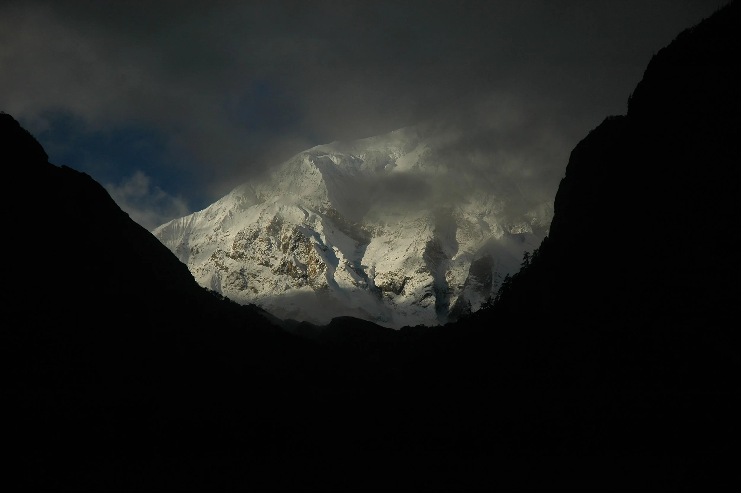 Snow-capped mountain peak seen through dark mountain valleys in a cloudy sky.