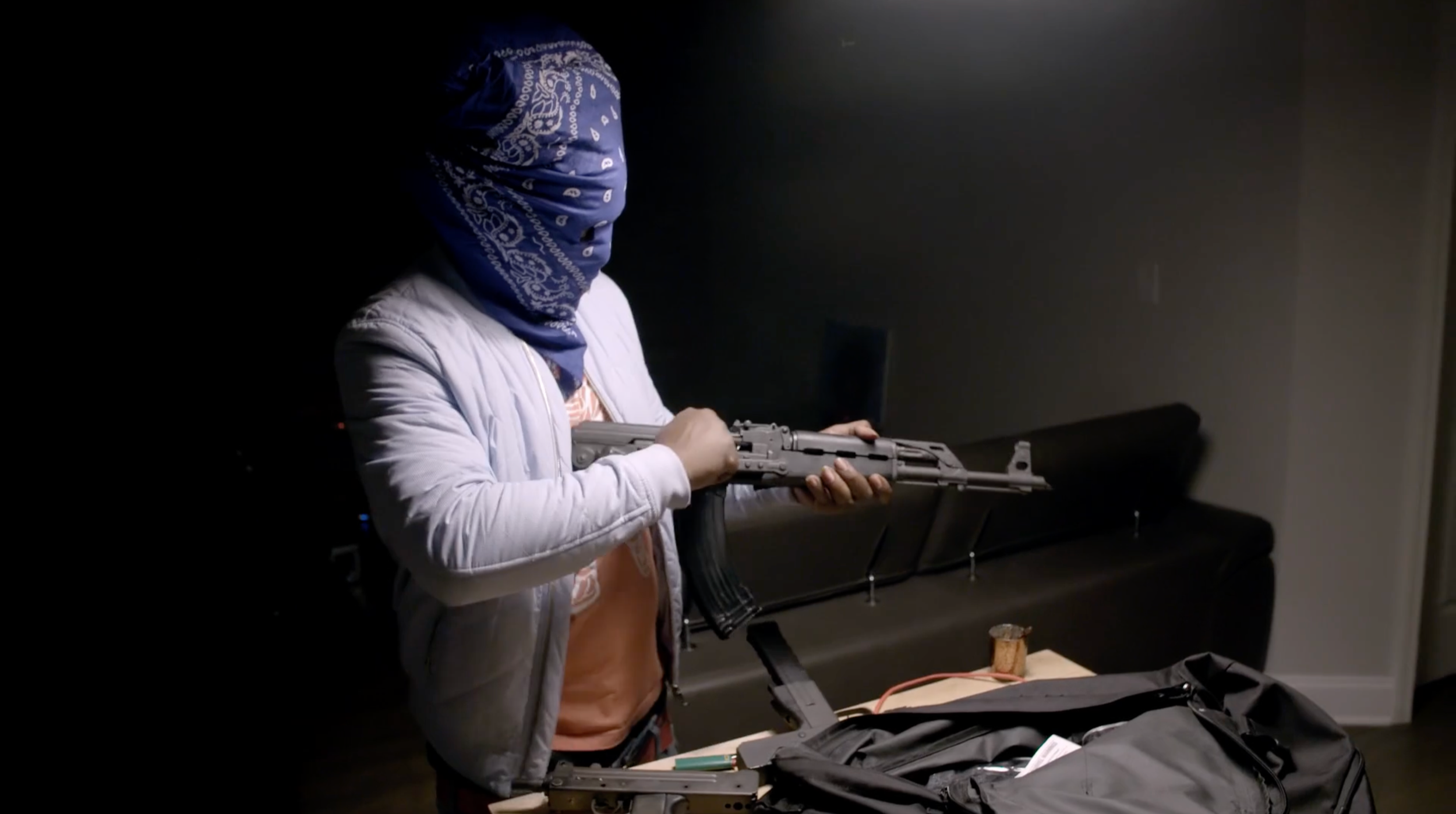 Person wearing a blue bandana over their face, holding a firearm in a room with a black couch and various objects on a table.
