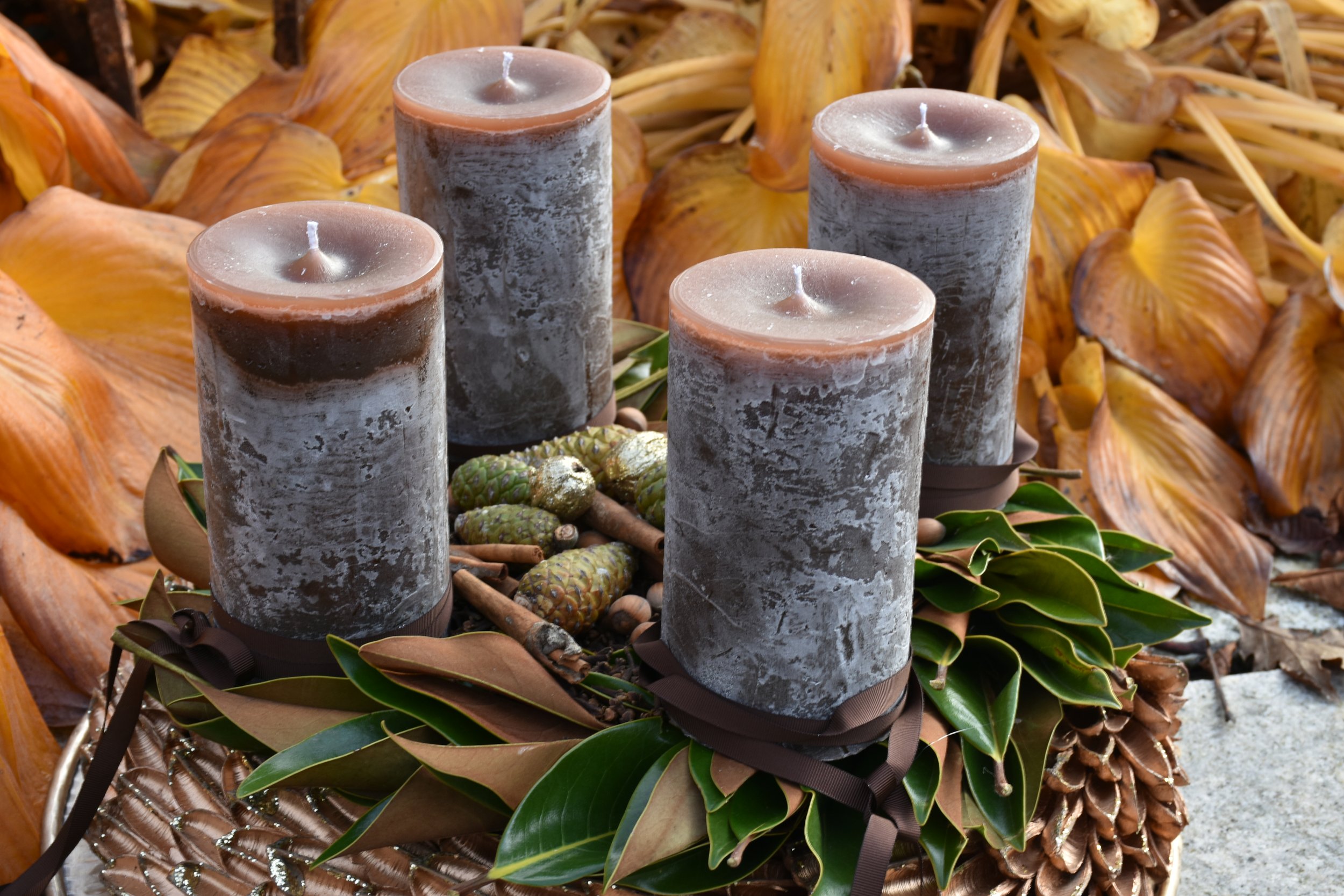 Vier graue Kerzen auf einem dekorativen Korb, umgeben von Blättern und getrockneten Früchten, mit herbstlichen Dekorationen im Hintergrund.
