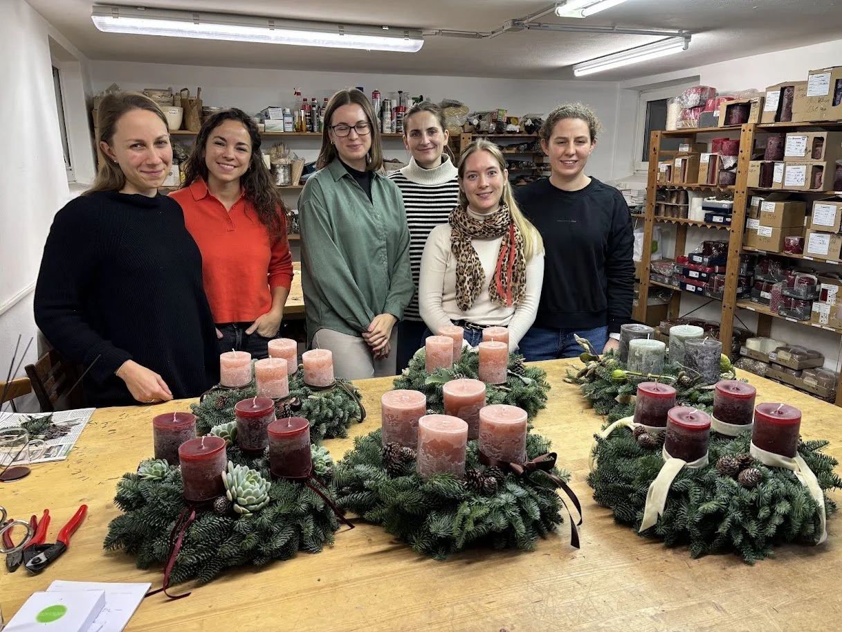 Sechs Frauen stehen hinter einem Tisch mit vier Adventskränzen aus Tannengrün, roten und rosa Kerzen, in einem Raum mit Holzregalen, die andere Dekorationen und Bastelmaterialien aufbewahren.