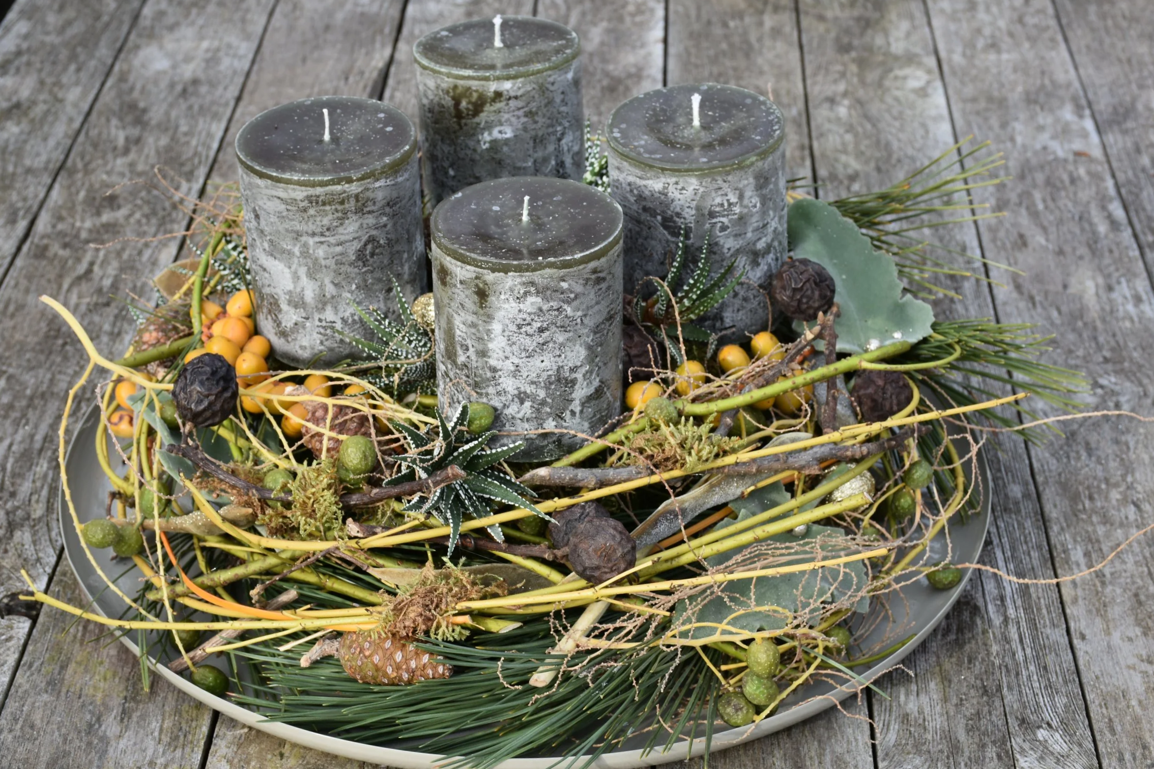 Dekorativer Adventskranz mit vier schwarzen Kerzen, grünen Zweigen, kleinen gelben Beeren, Tannenzapfen und weiteren Naturmaterialien, auf einer Holztischoberfläche.