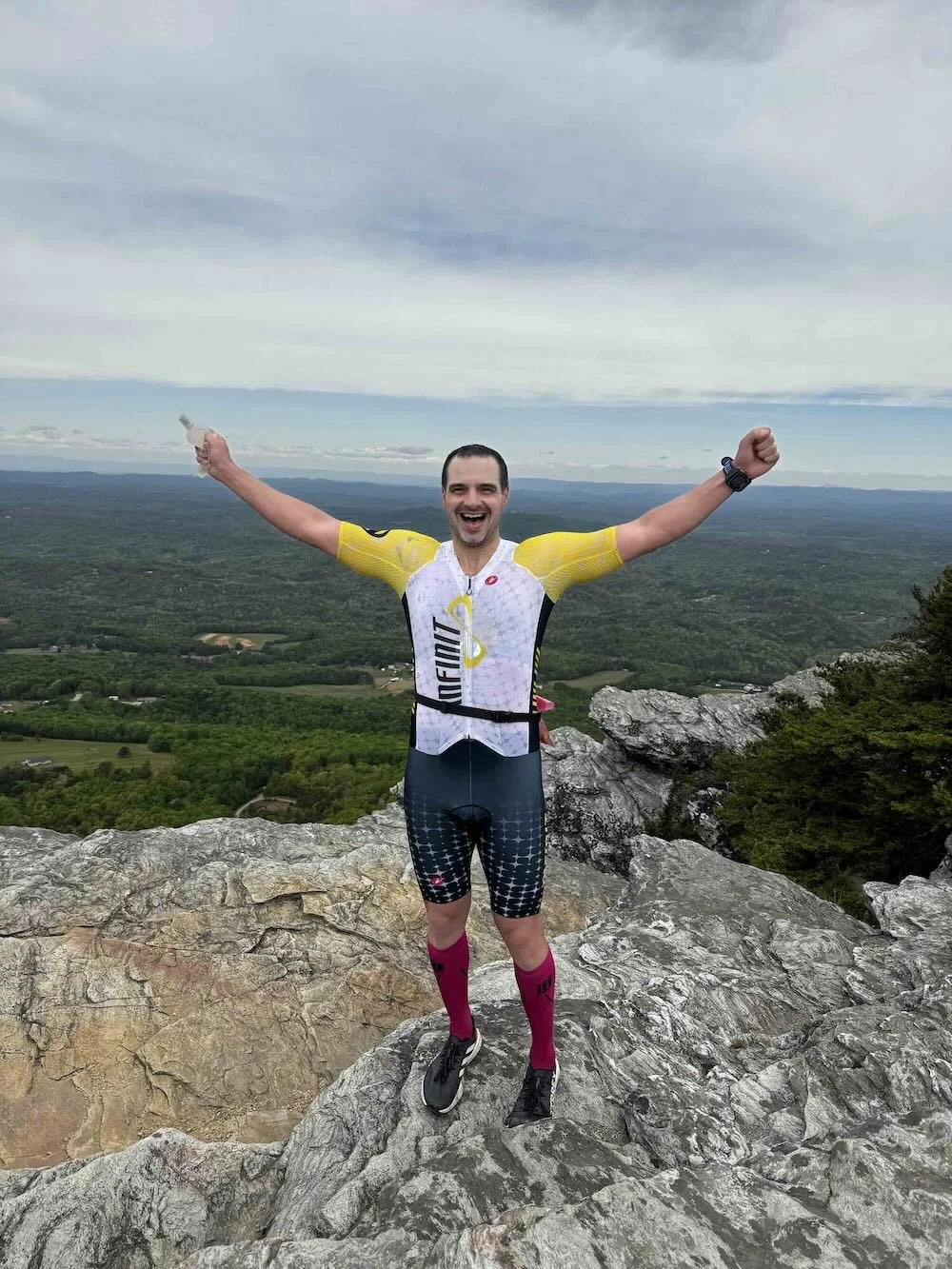 A man in cycling gear standing on a mountain rock with his arms raised in victory, overlooking a expansive green landscape with a cloudy sky.