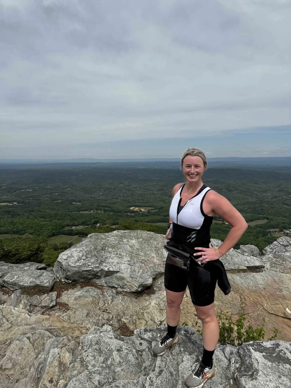 Woman in athletic gear standing on rocky ledge with scenic green landscape and cloudy sky in the background.