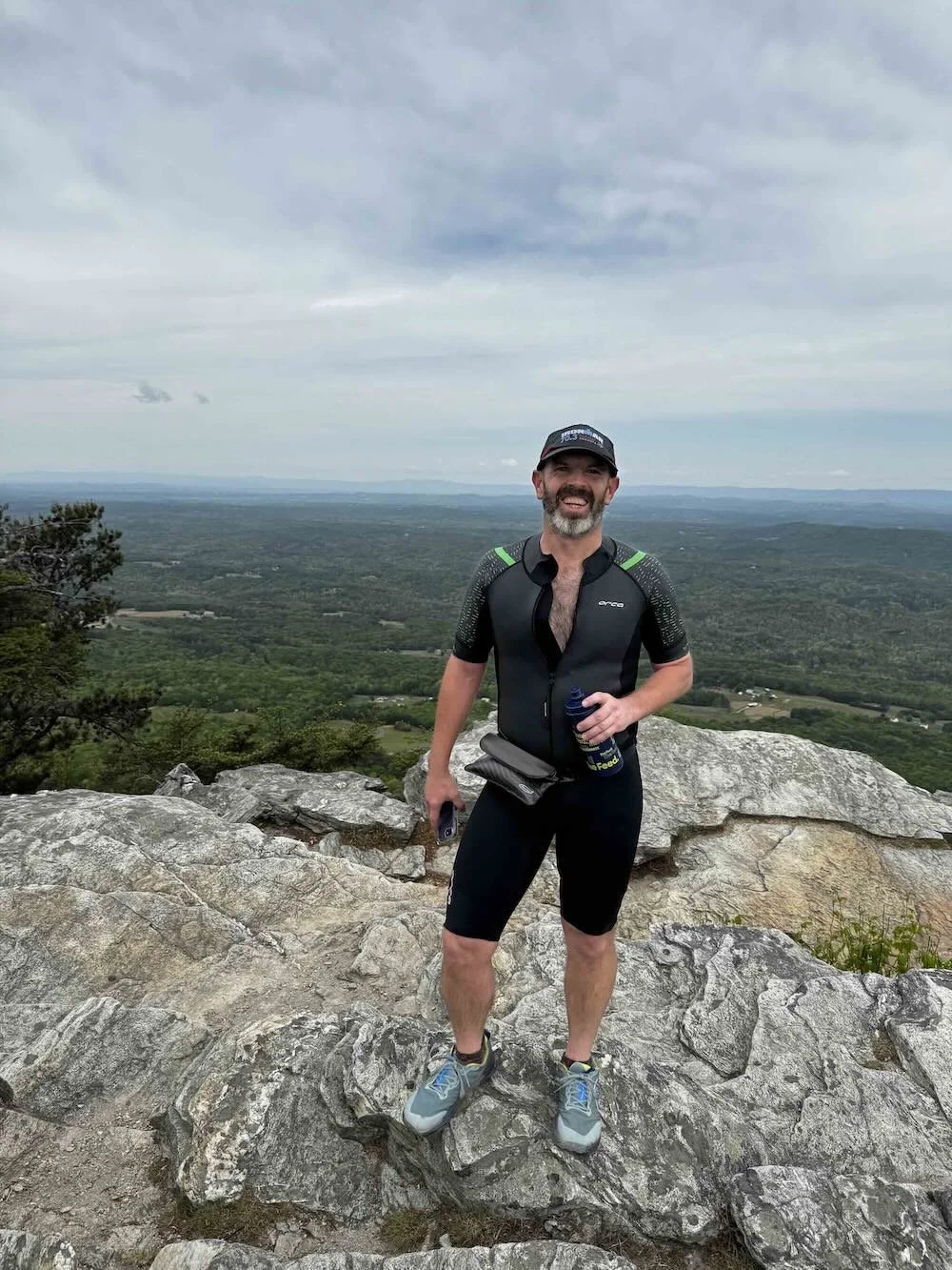 A man in athletic gear standing on rocky top of a mountain with a wide expanse of green landscape and cloudy sky in the background.
