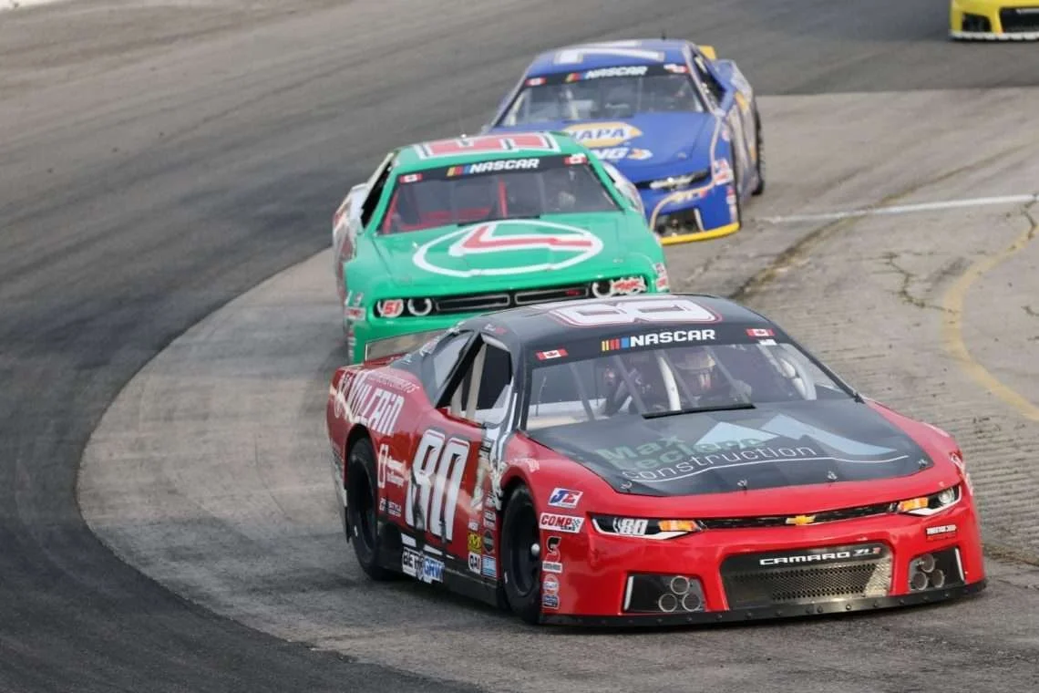 Three NASCAR stock cars race around a track, led by a red No. 80 Chevrolet Camaro, followed by green and blue cars.