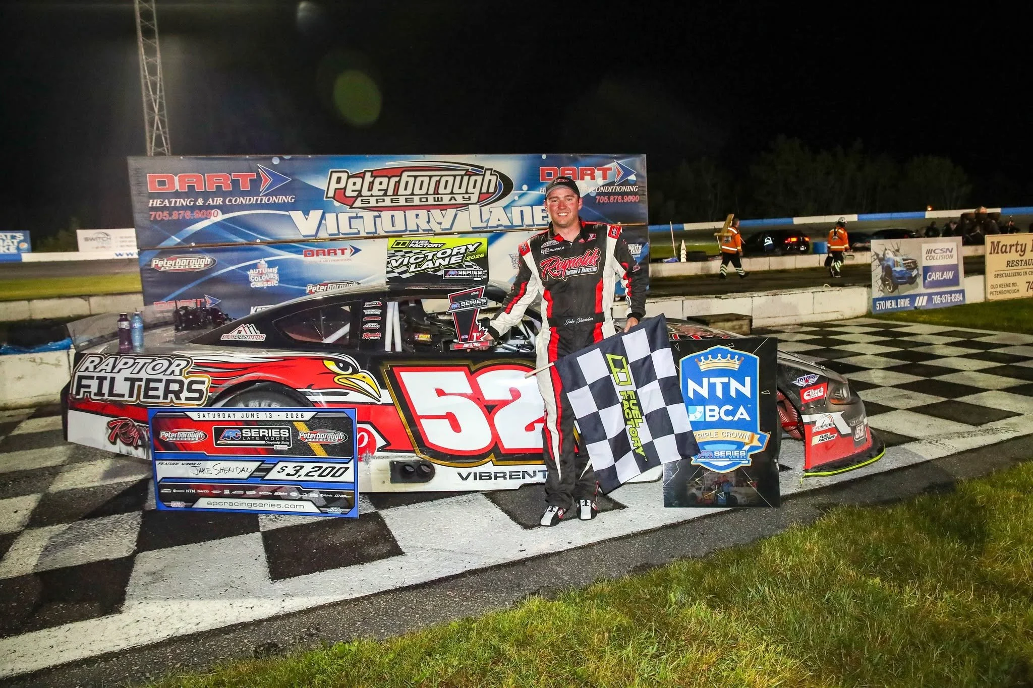 Race car driver Jake Sheldon poses with a checkered flag, trophy, and No. 52 stock car after winning at Peterborough Speedway.