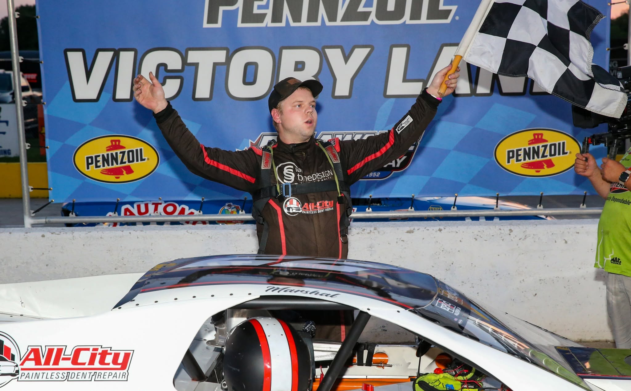 Race car driver celebrating with a checkered flag beside a stock car at Victory Lane