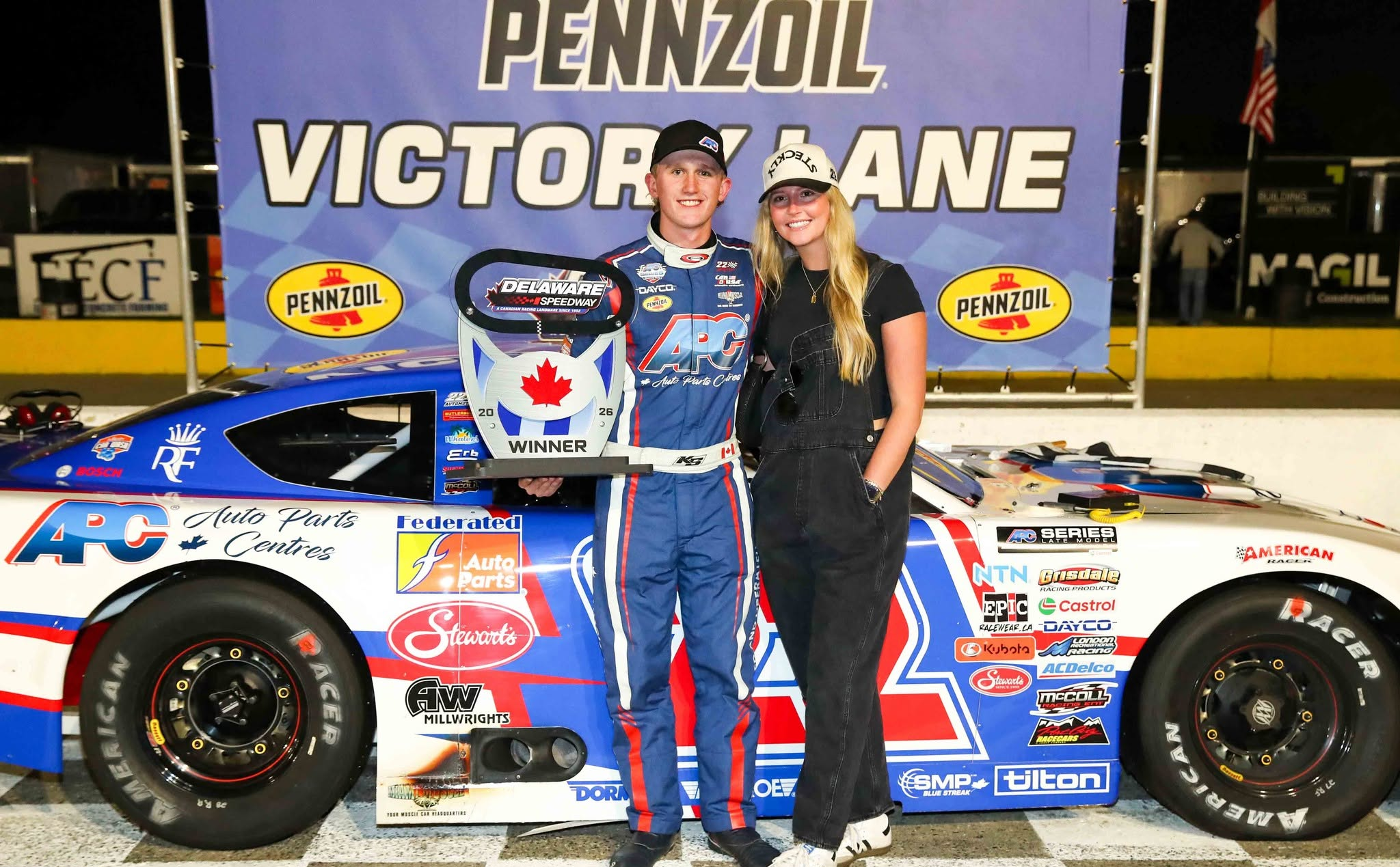 Race car driver and woman pose with a winner trophy beside a sponsored stock car at Delaware Speedway.