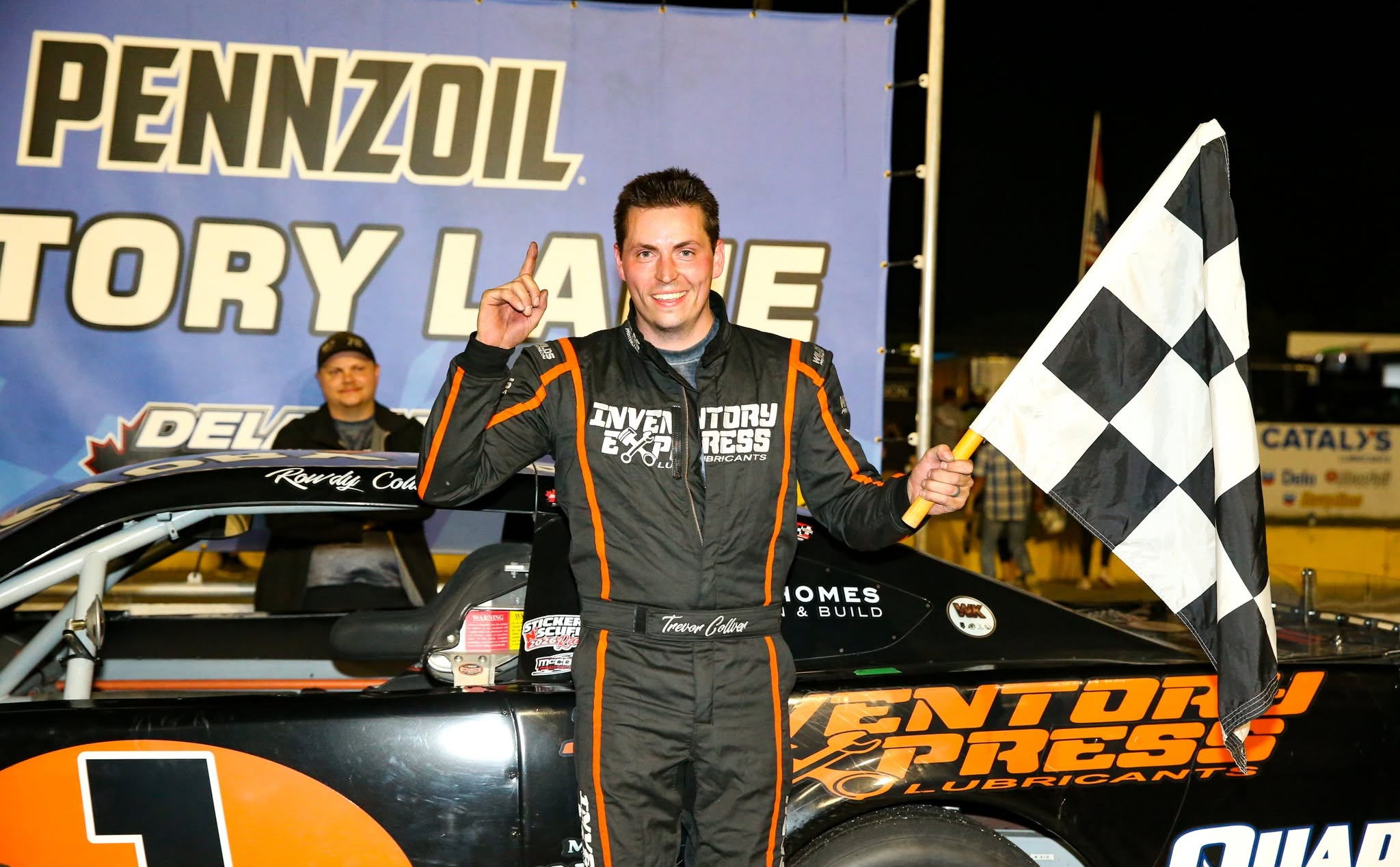 Smiling race car driver Trevor Collver holds a checkered flag in front of a sprint car at a Pennzoil Victory Lane celebration.