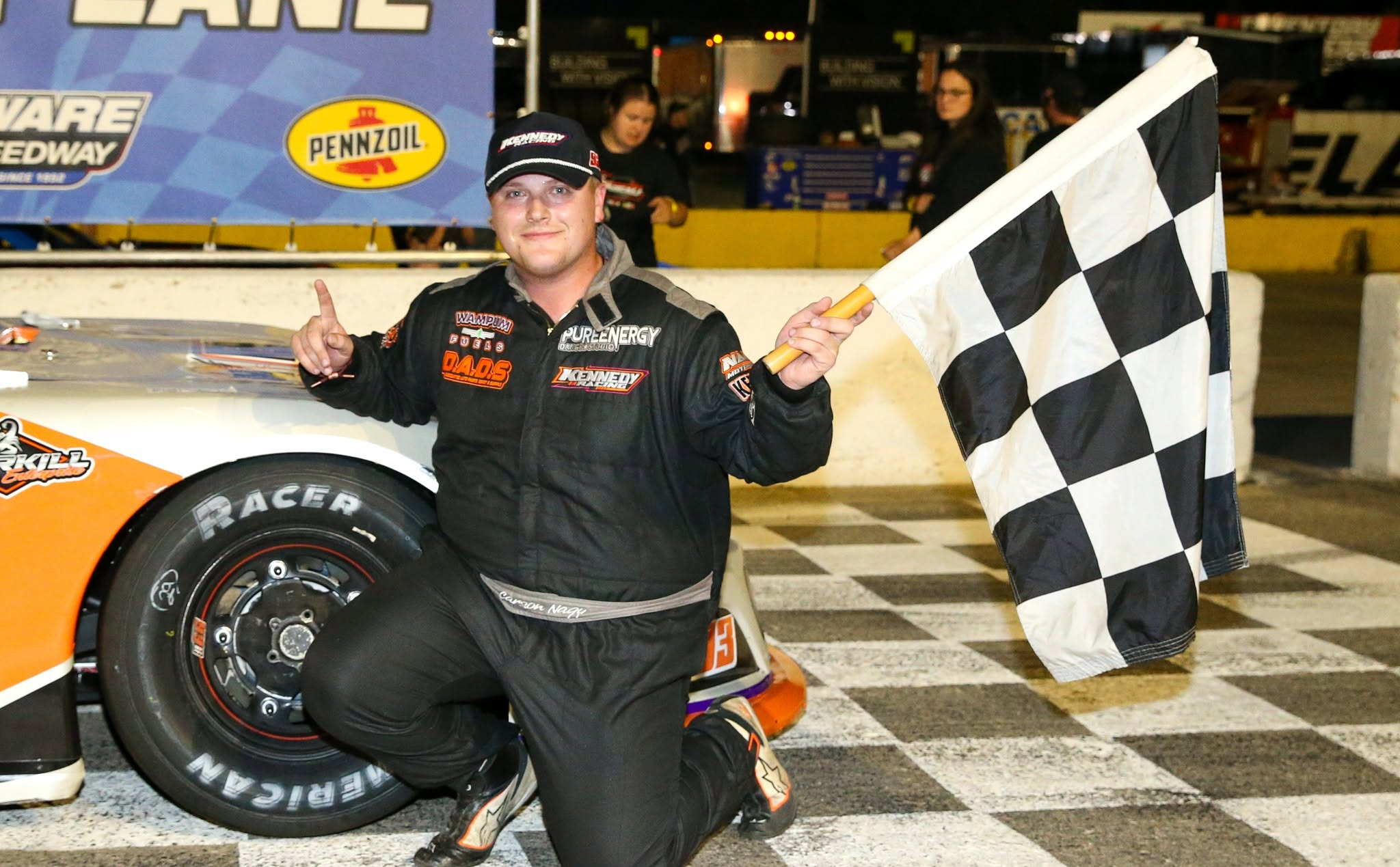 Race car driver kneeling beside a stock car and holding a checkered flag after a victory at the track