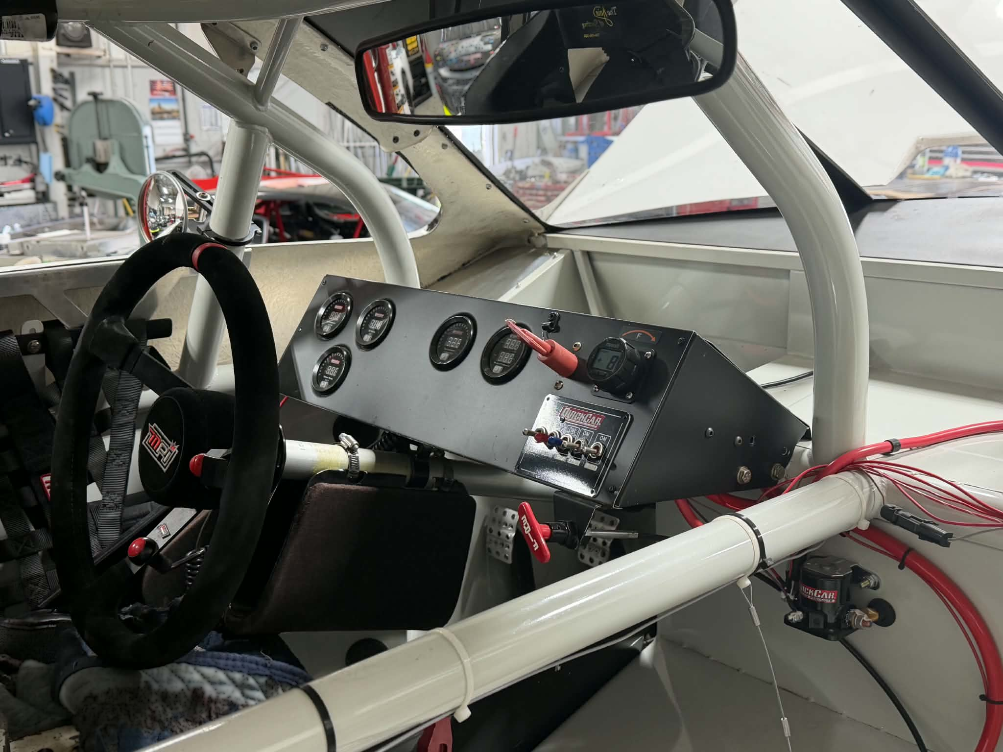 Interior of a race car showing a roll cage, racing steering wheel, dashboard gauges, switches, and fire suppression equipment.