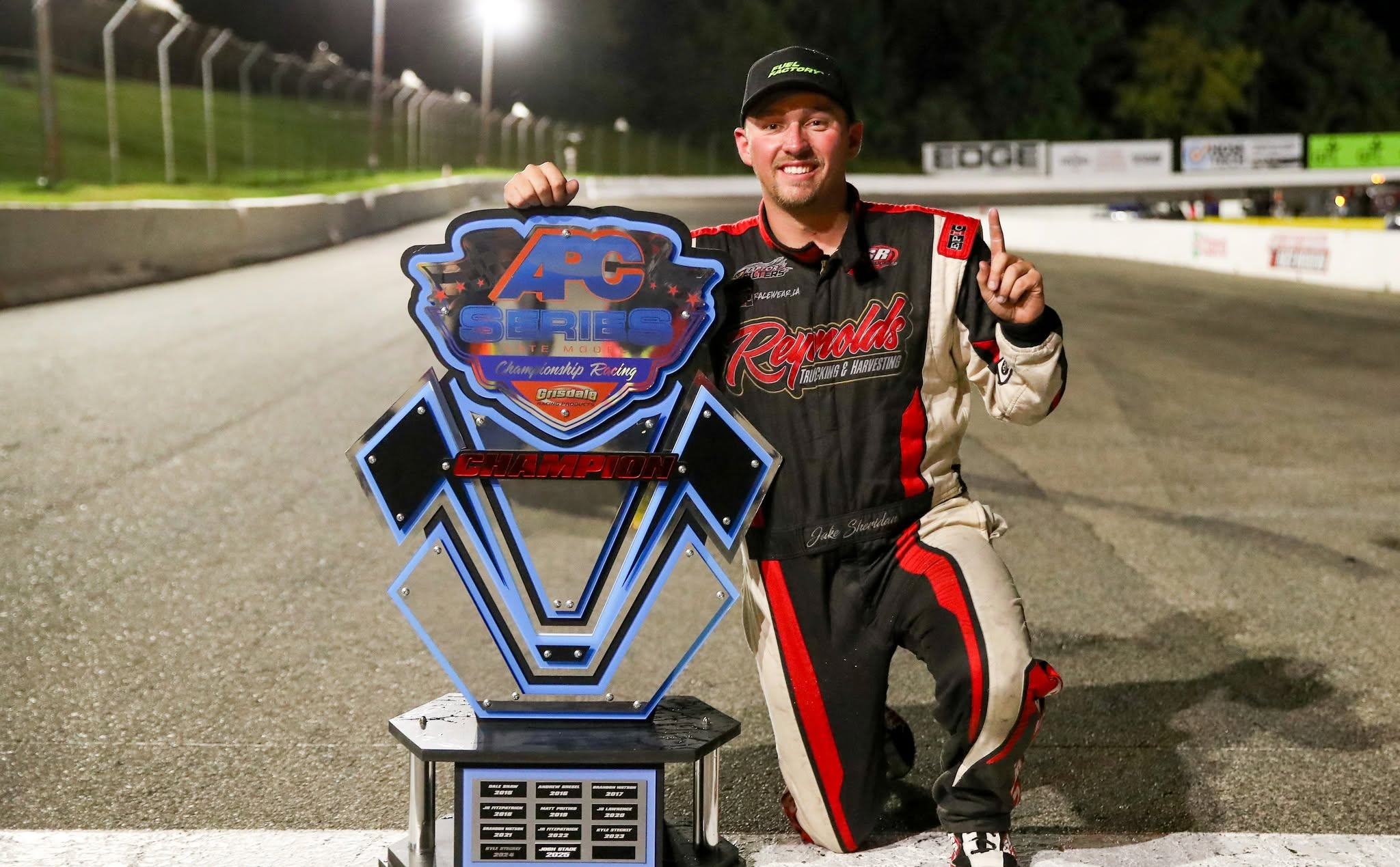 Jake Sheridan driver kneeling beside a large APC Series championship trophy on a nighttime delaware speedway