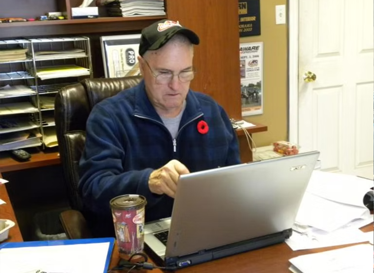 Man wearing a baseball cap and glasses working on a laptop at an office desk