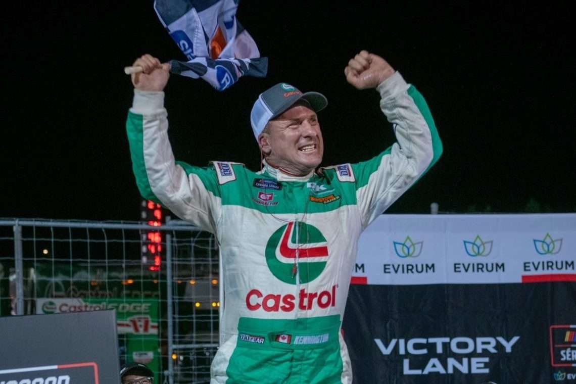 DJ Kennington Race car driver in a green Castrol racing suit celebrates victory with raised fists and a checkered flag at night