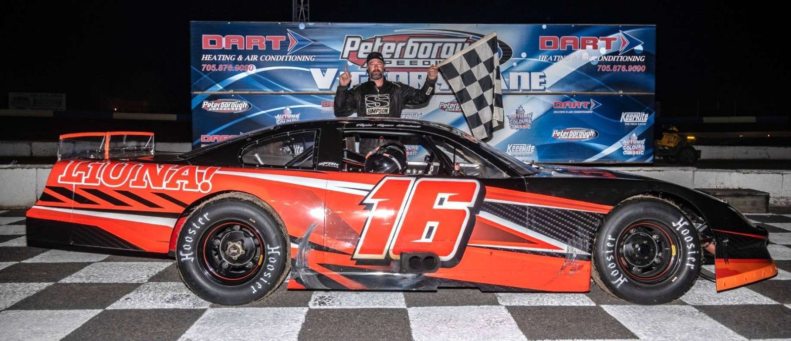 Red and black number 16 stock car parked on a checkered victory lane as the driver holds a checkered flag.