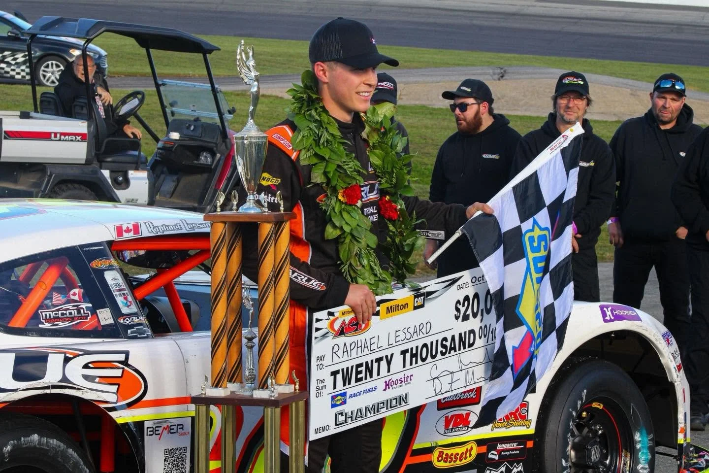 Racing driver Raphael Lessard celebrates a ACT Championship with a trophy, oversized $20,000 winner’s check, Canadian flag, and laurel wreath beside his race truck.