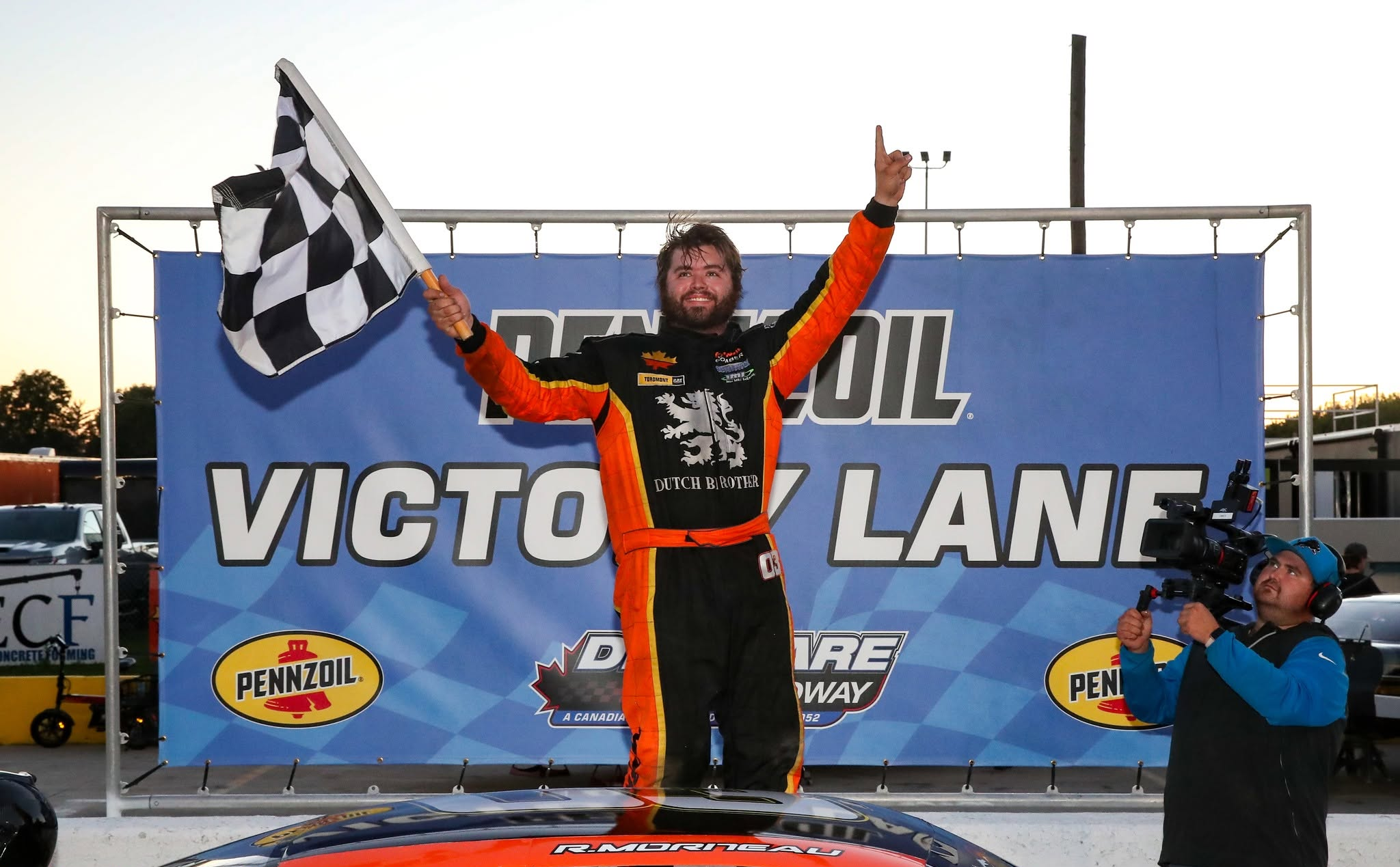 Smiling race car driver in an orange-and-black suit celebrates with a checkered flag in front of a victory lane sponsor banner.