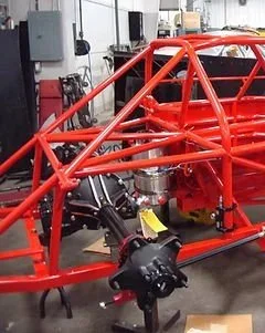 Bright red tubular steel race car chassis with exposed suspension and drivetrain components in a workshop