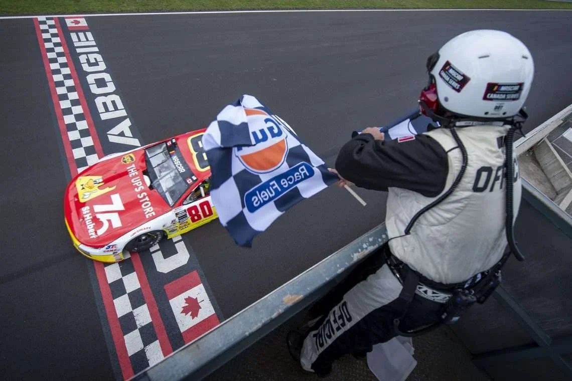 Race official waves a checkered flag over a red No. 80 stock car crossing the Calabogie finish line.