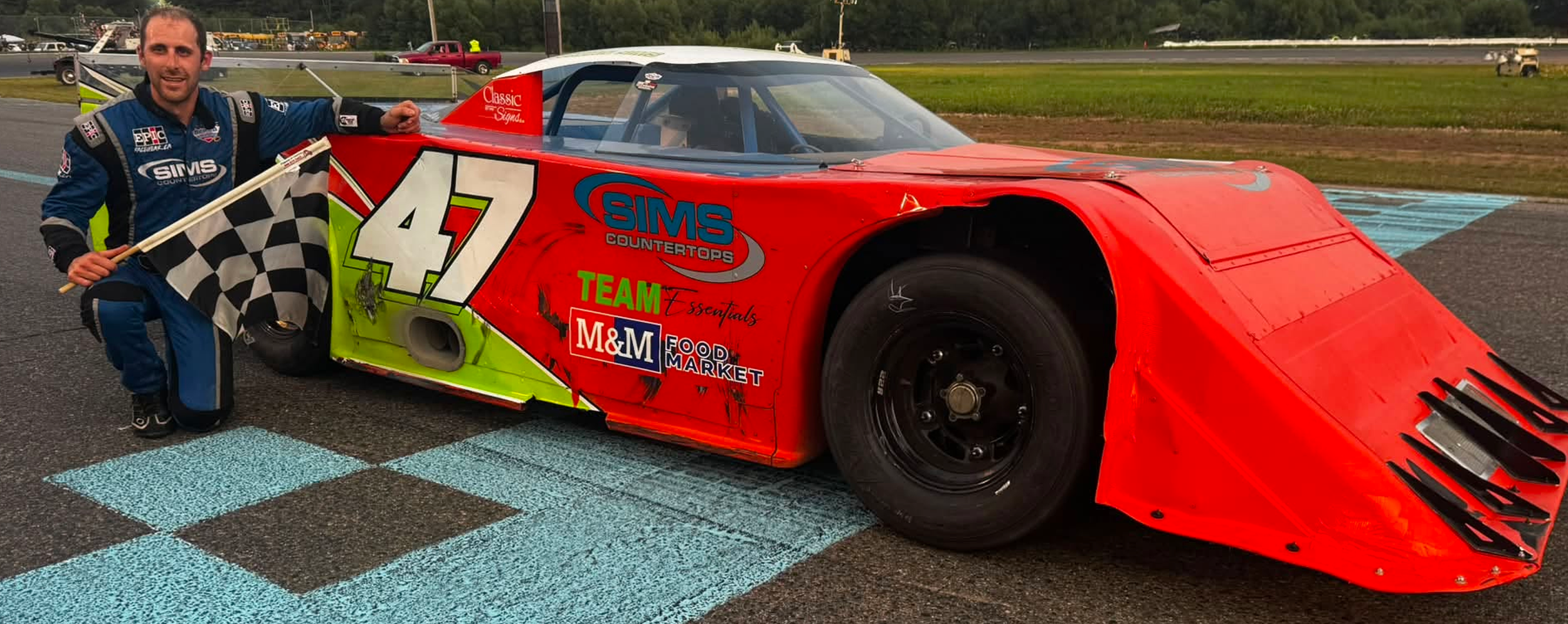 Race car driver holding a checkered flag beside a red No. 47 stock car on a racetrack