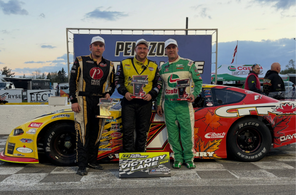 Three race car drivers pose with trophies beside a colorful stock car on a racetrack victory lane.