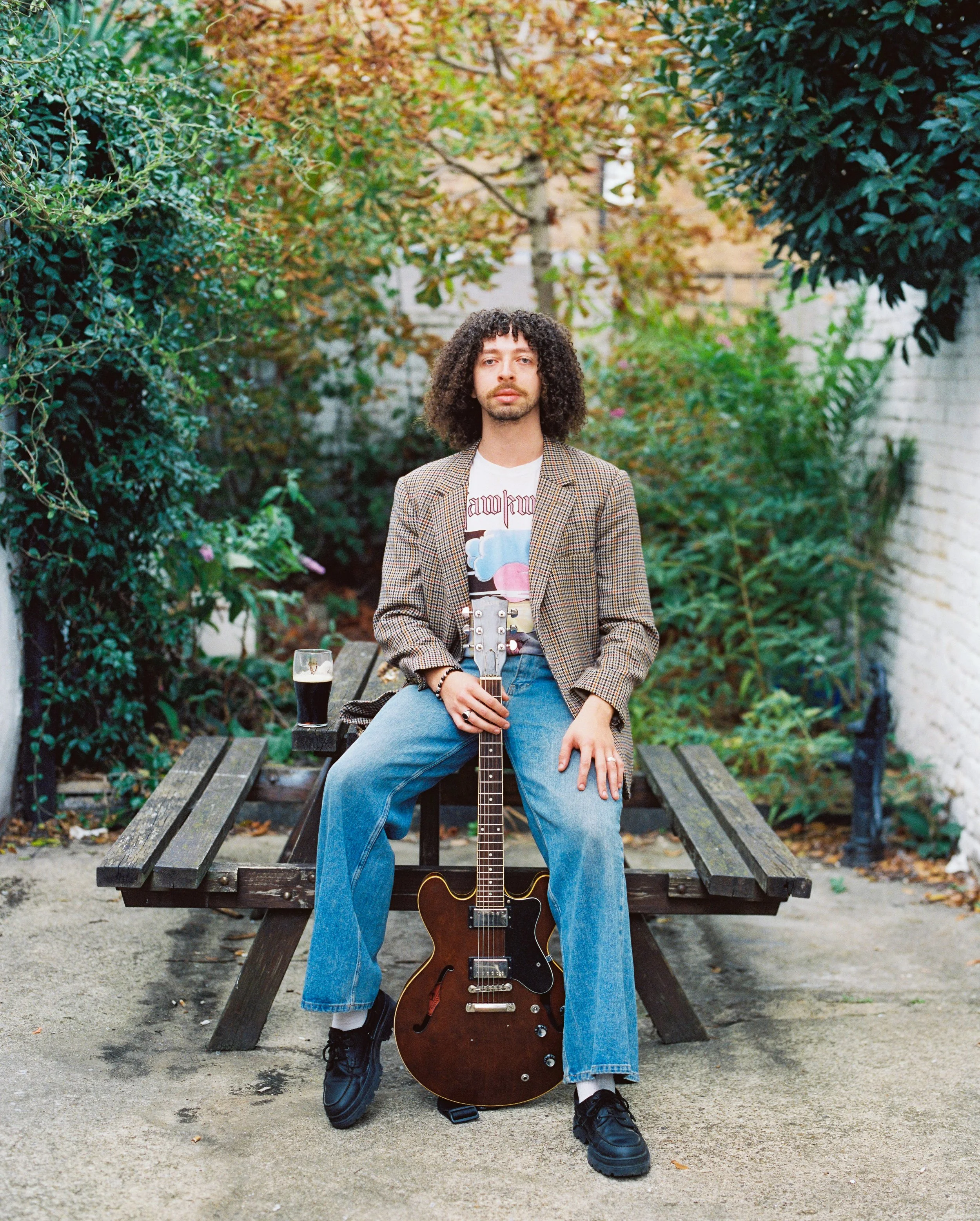 A person with curly hair and light skin sitting on a park bench, holding a guitar, with a glass of dark beverage on the bench beside them, surrounded by trees and greenery