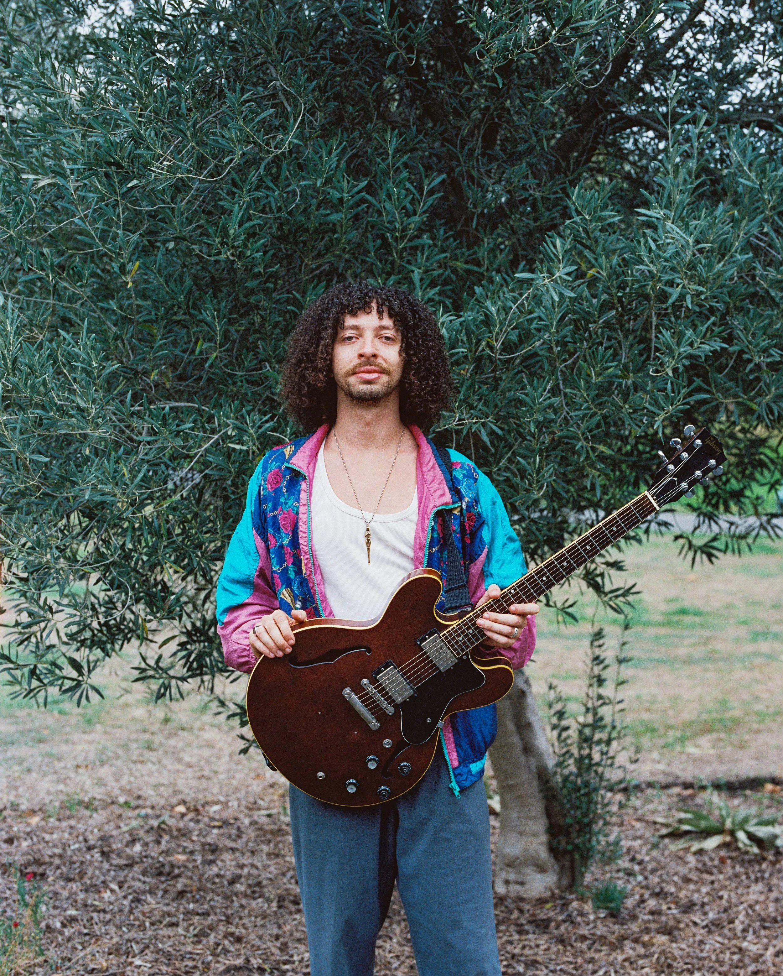 A person standing outdoors in front of a large bush, holding a guitar. They have curly hair, wearing a colorful jacket over a white shirt, and blue jeans.