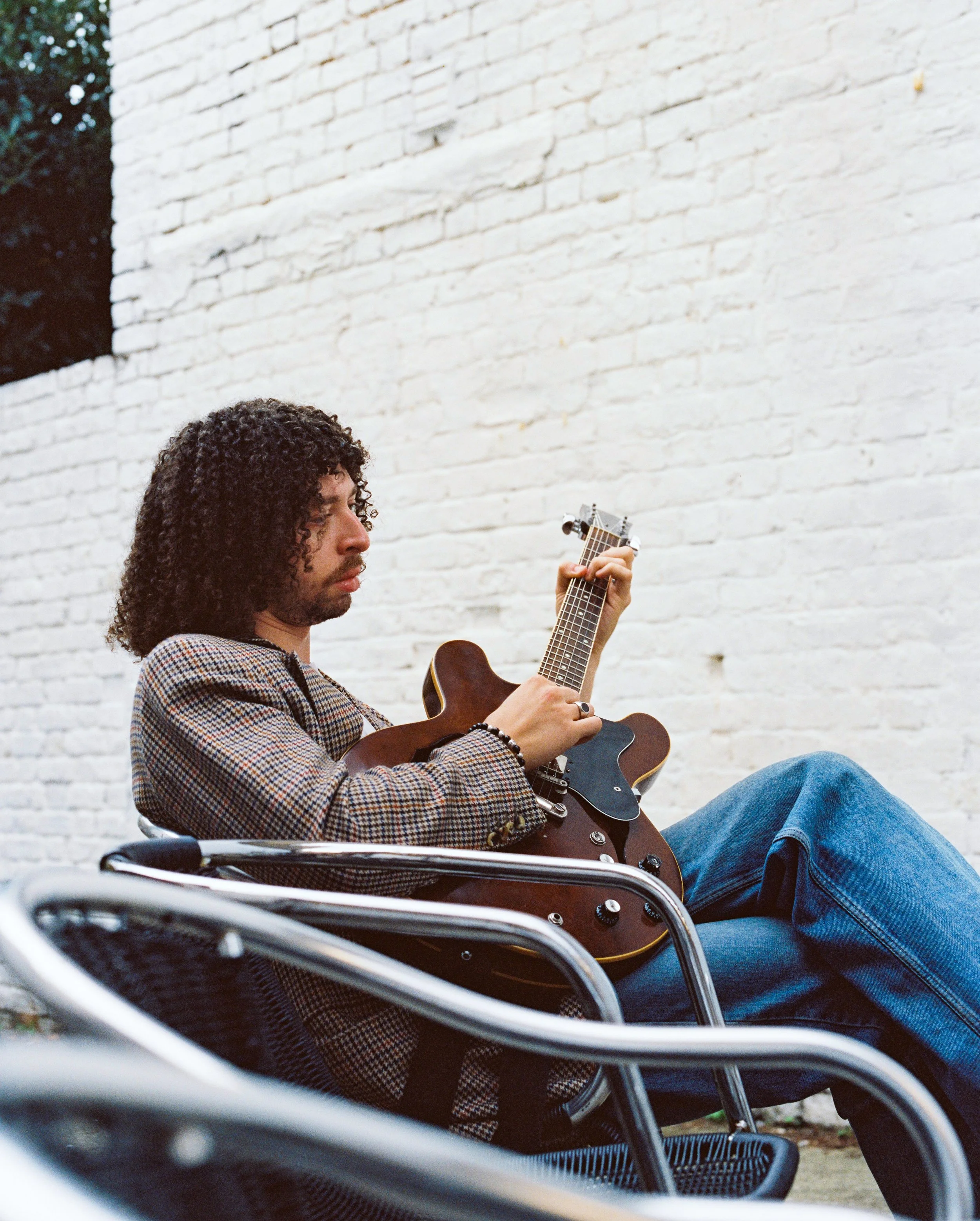 A man with curly dark hair playing an acoustic guitar while sitting outside against a white brick wall.