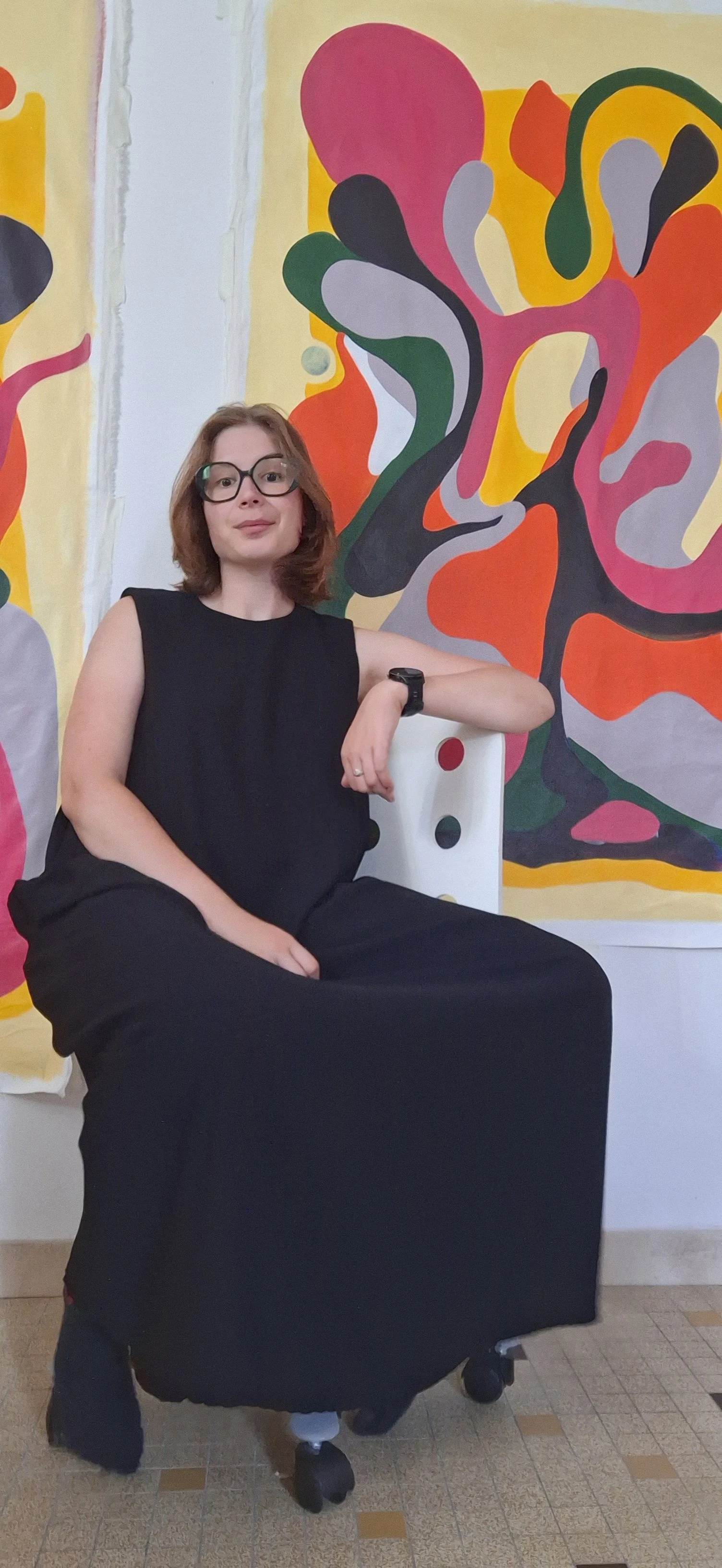 Femme portant une tenue noire et des lunettes, assise devant une fresque abstraite colorée.