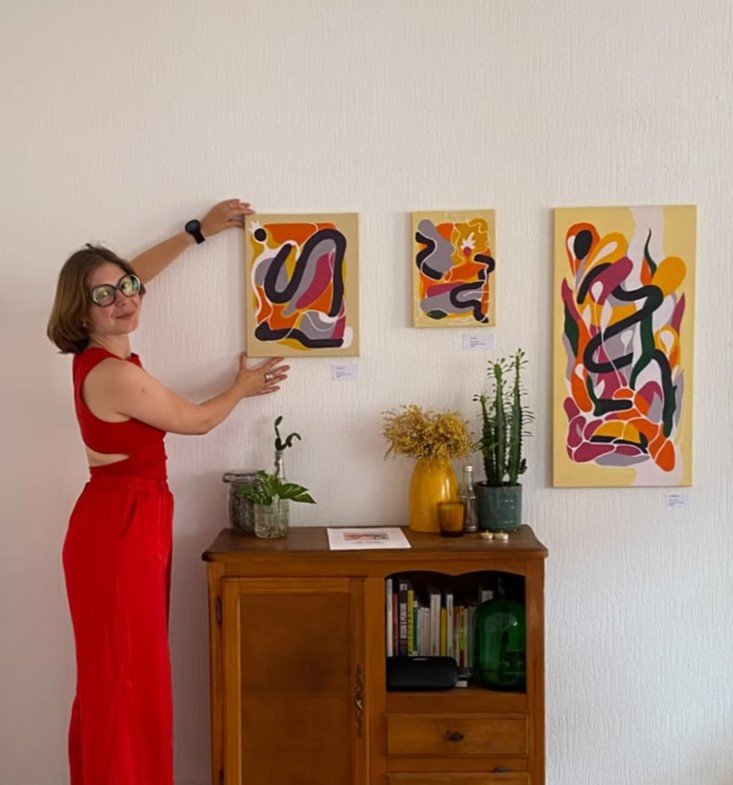 Femme en robe rouge présentant trois tableaux abstraits colorés au-dessus d’un buffet décoré de plantes et de fleurs.