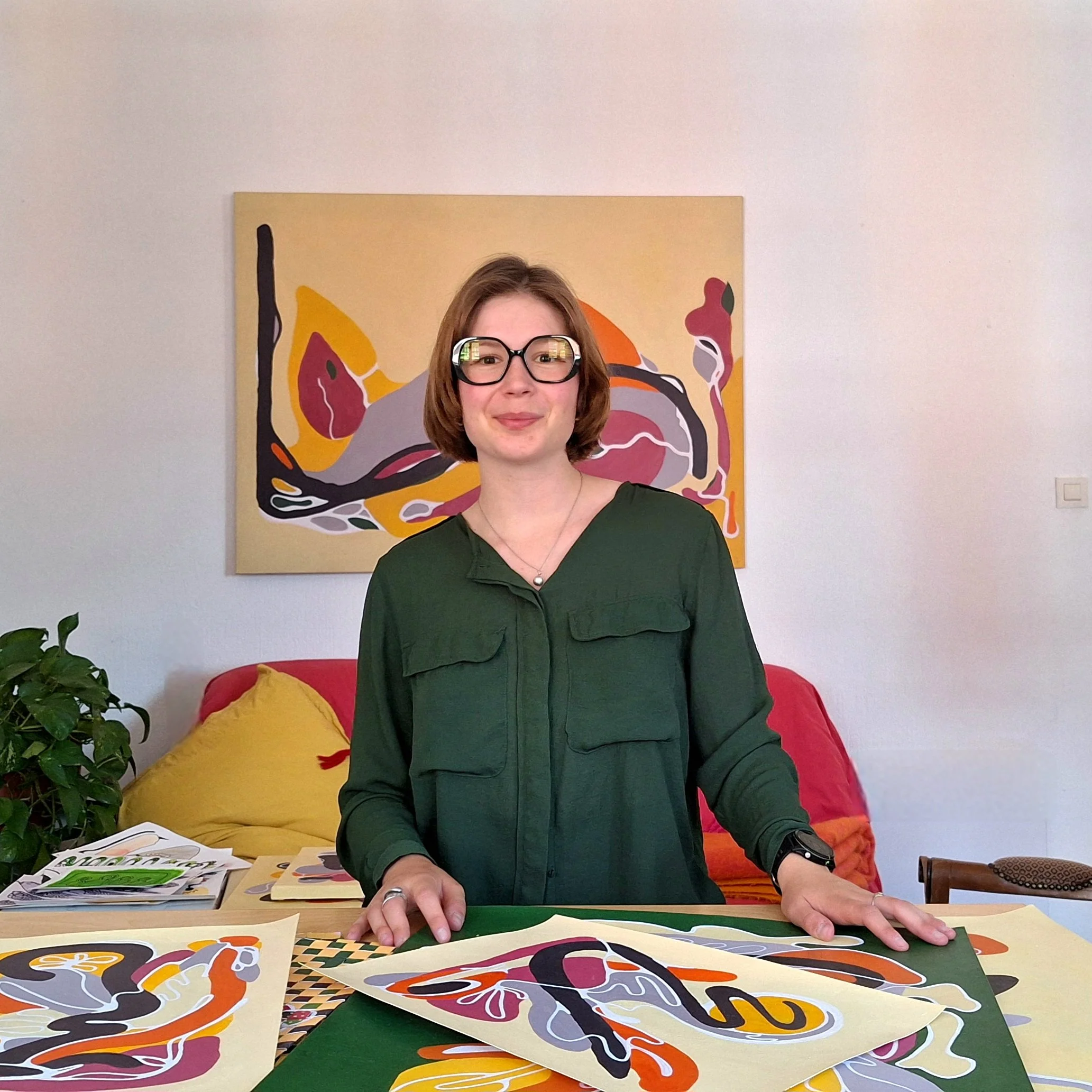 Artiste portant des lunettes devant ses peintures abstraites colorées, assise à une table dans un intérieur chaleureux.