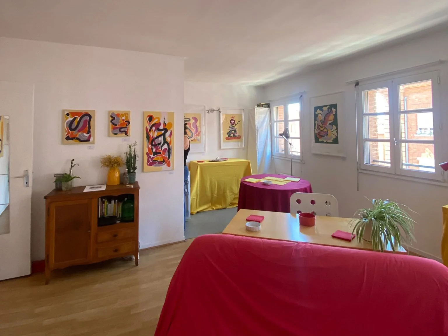 Intérieur lumineux d’une galerie d’art avec peintures abstraites colorées, tables dressées et canapé rouge.