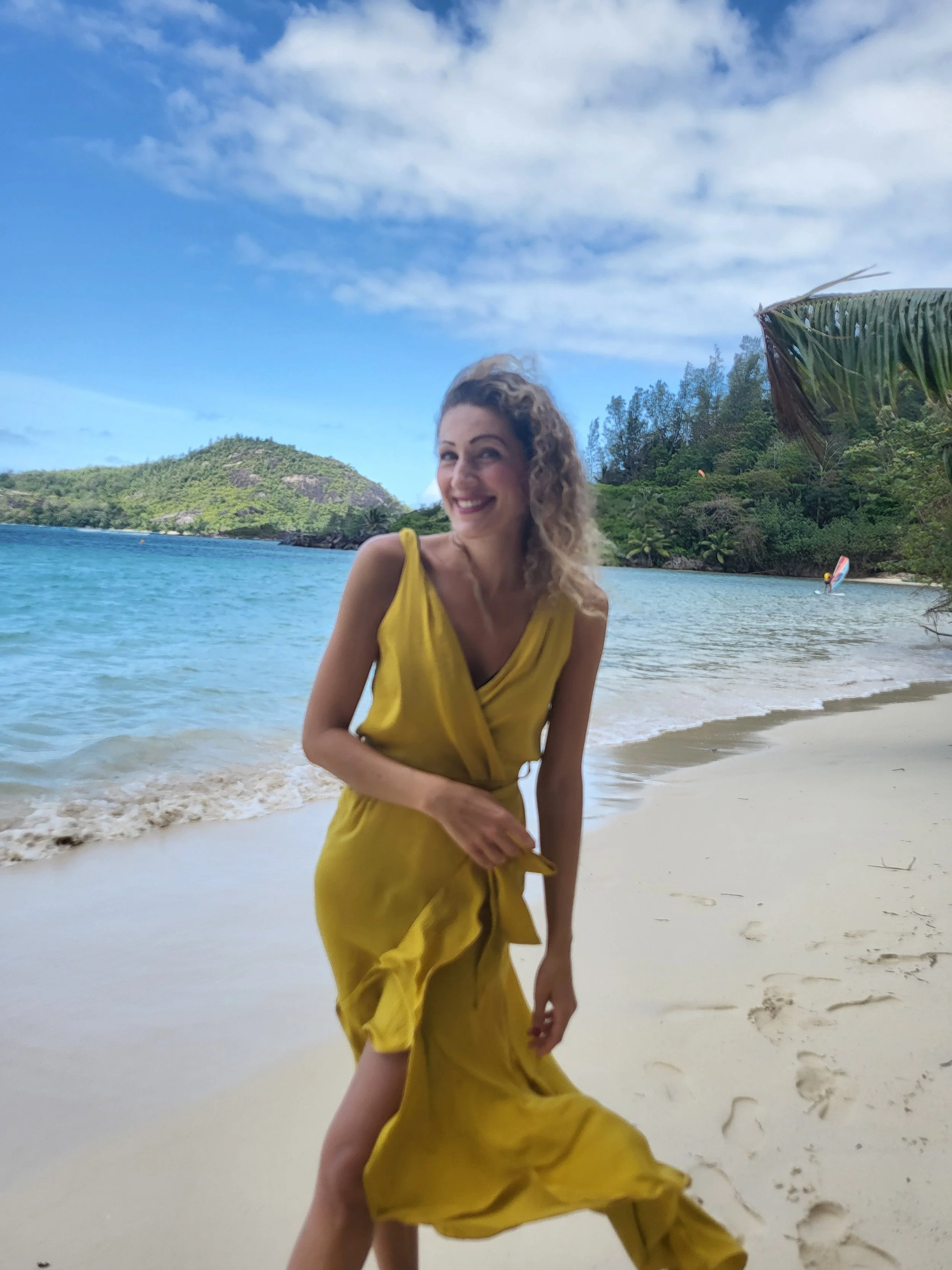 A woman with curly hair wearing a yellow dress is smiling and standing on a sandy beach near the water, with a lush green island and blue sky in the background.