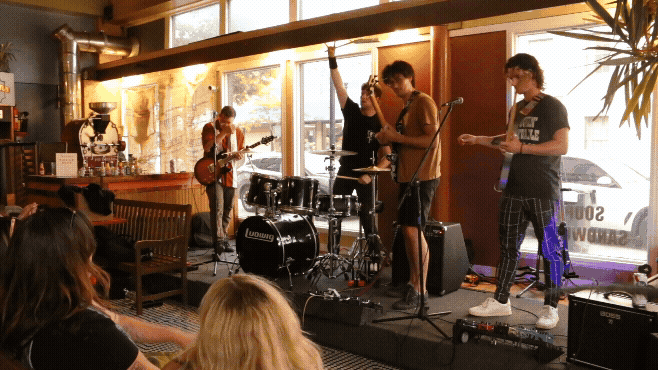 A band performing live music on stage with guitar, drums, and vocals inside a cozy venue with large windows and audience watching.