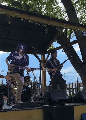 Two musicians playing electric guitars on an outdoor stage during daytime, with a drum set and amplifiers behind them, and a wooden fence and trees in the background.