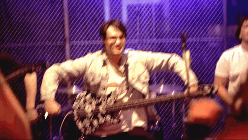 A person holding a guitar in a room with purple lighting and a chain-link fence background, smiling and pointing towards the camera.