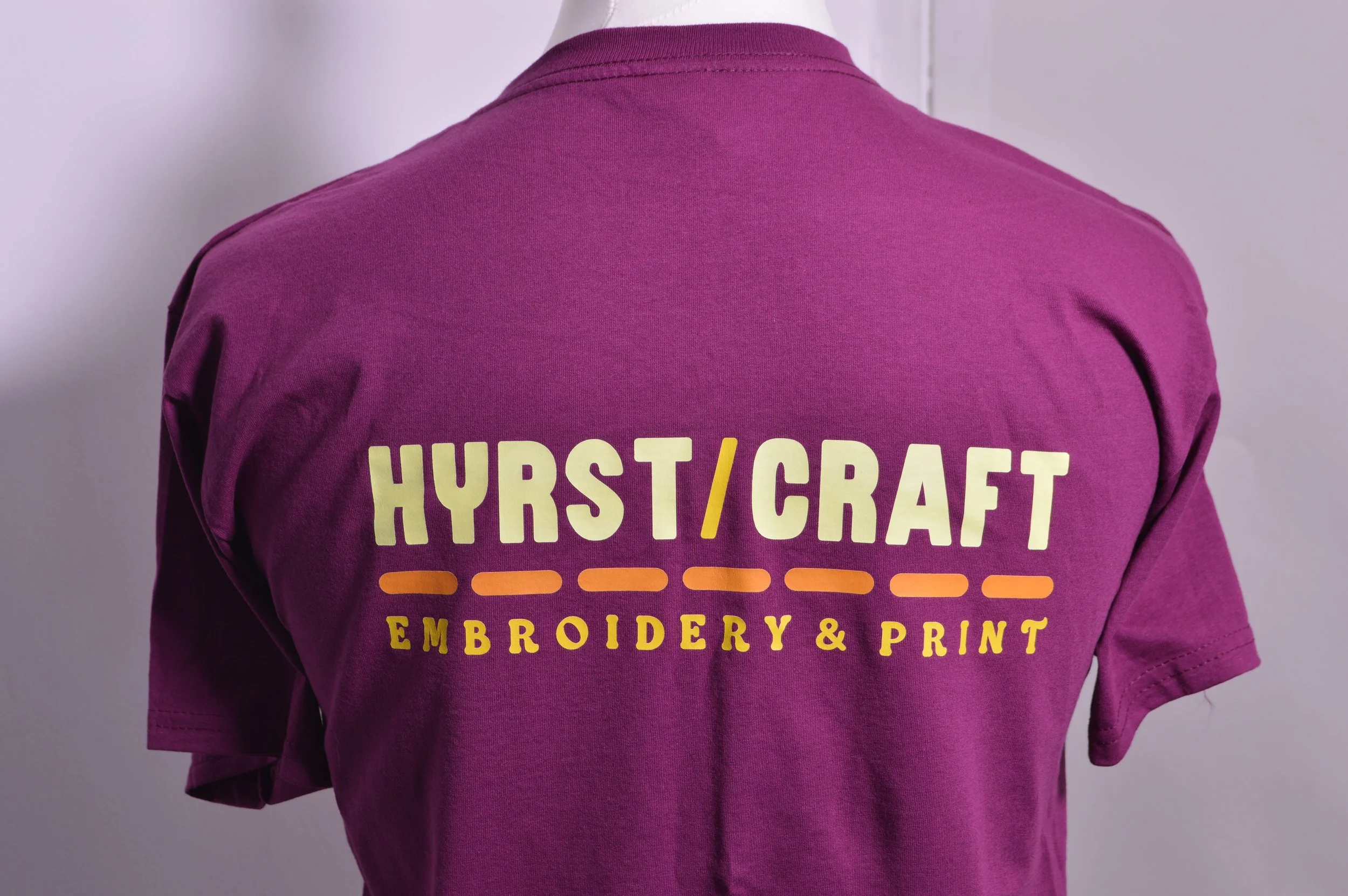 Back view of a person in a purple shirt with yellow and orange printed text about embroidery and print.