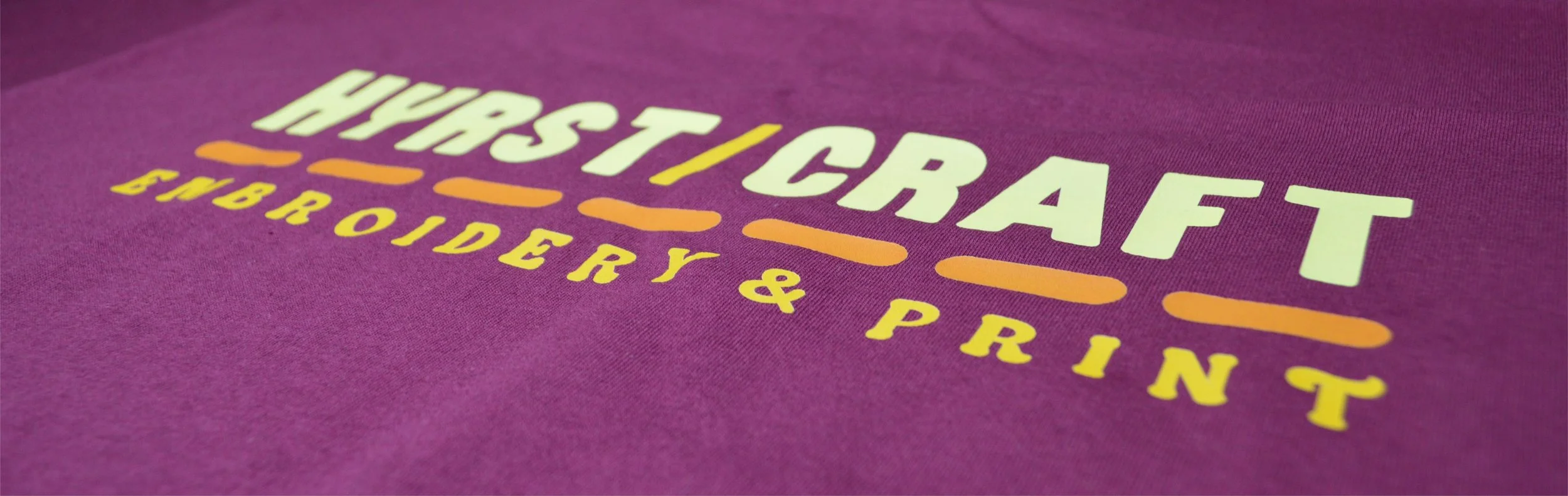 Close-up of a maroon T-shirt with yellow and white text that reads 'HYRSTCRAFT EMBROIDERY & PRINT'.