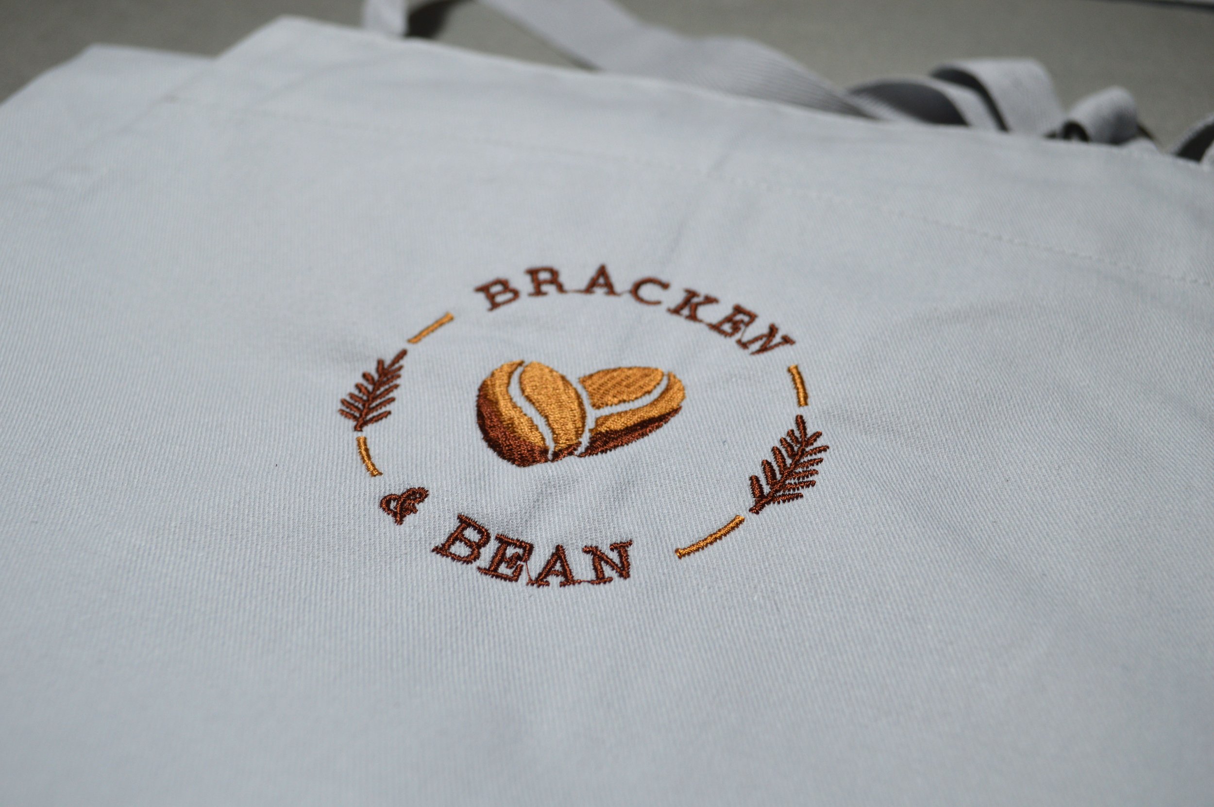 Close-up of embroidered logo on white fabric, featuring a coffee bean, the word 'BRACKEN,' and decorative leaves.
