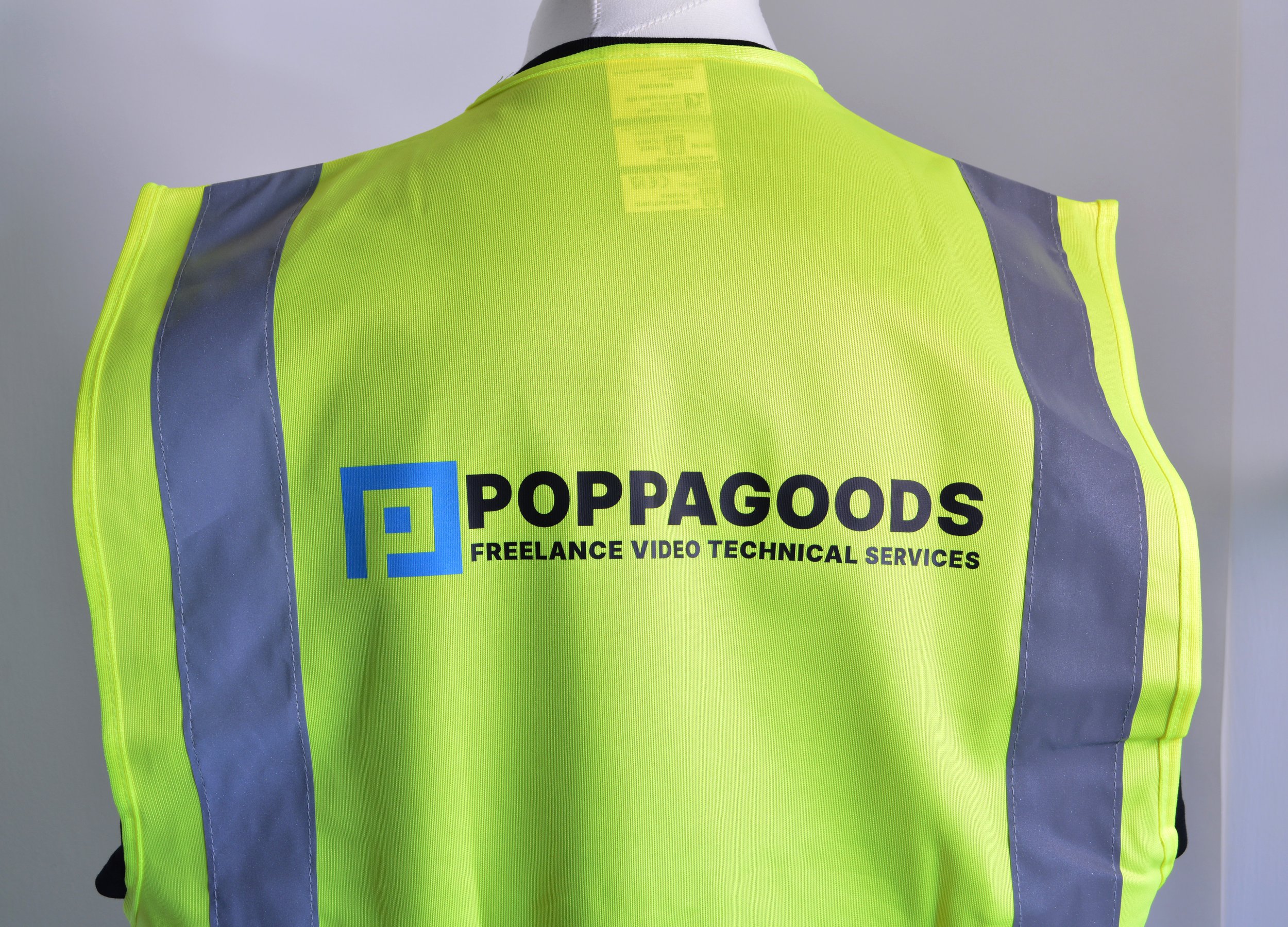 Back of a high-visibility yellow safety vest with gray reflective stripes and the logo of PoppGoods, a freelance video technical service company.