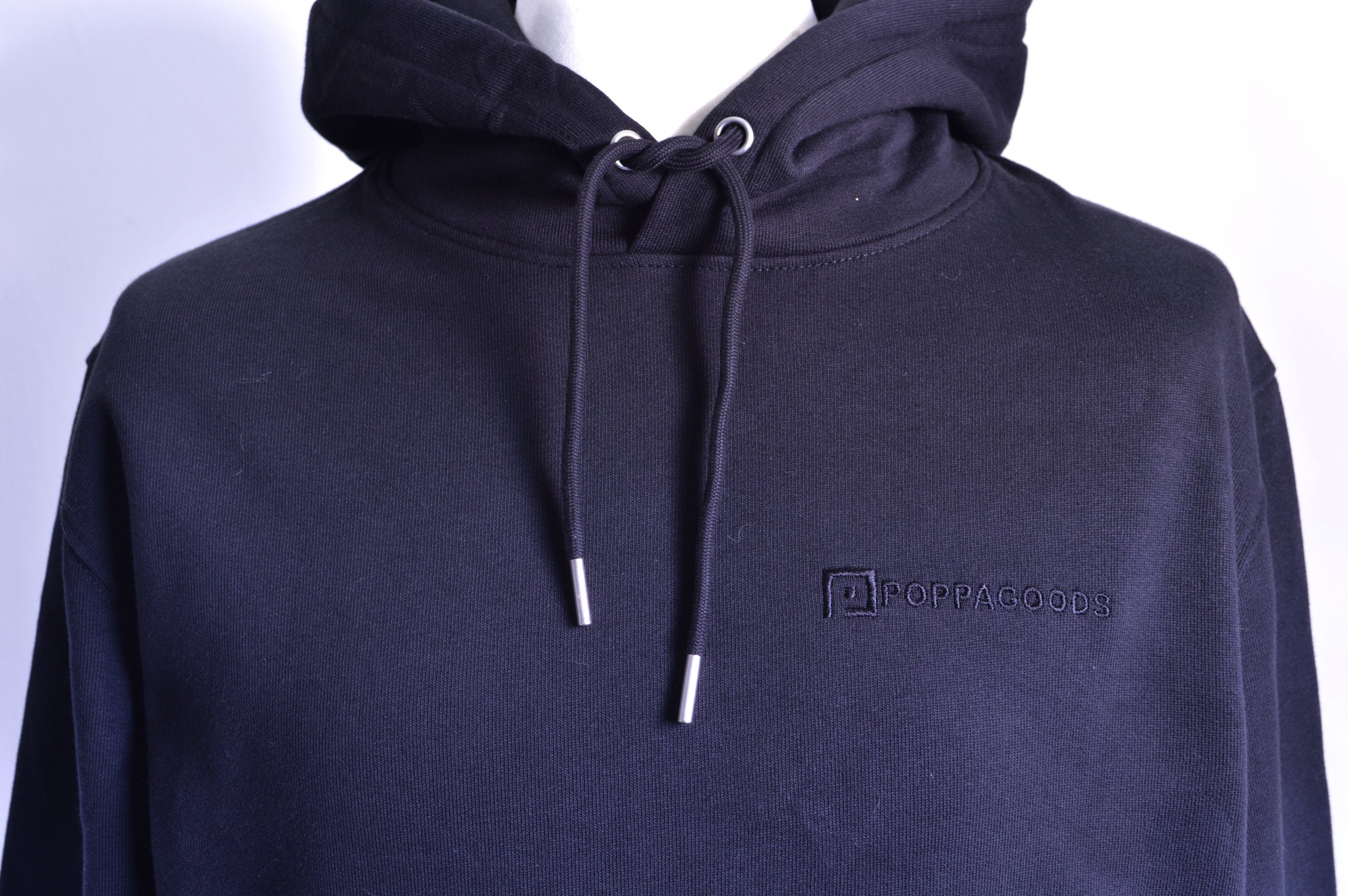 Close-up of a person wearing a black hoodie with a small embroidered logo of Poppagoods on the chest, with the hoodie strings hanging down and black embroidered logo on front left breast.
