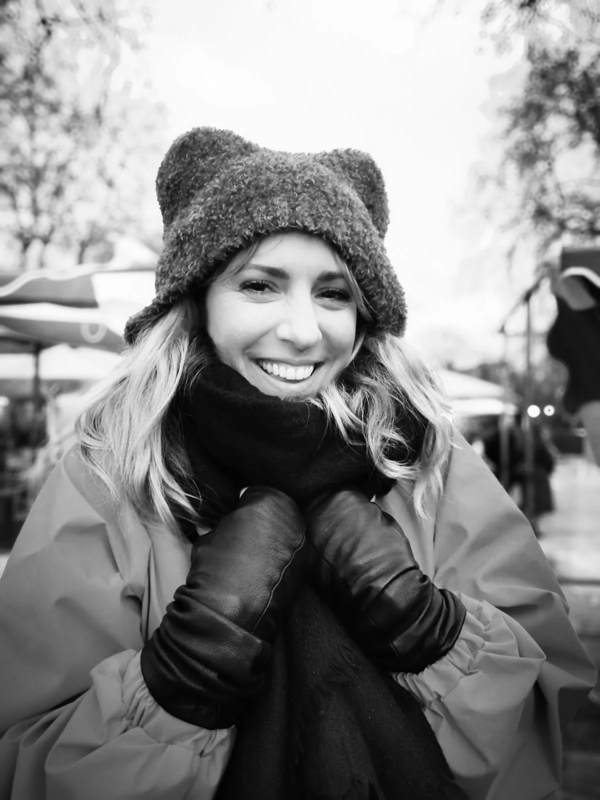 A woman smiling outdoors, wearing a warm fur hat, a scarf, gloves, and a coat, in a black-and-white photo.