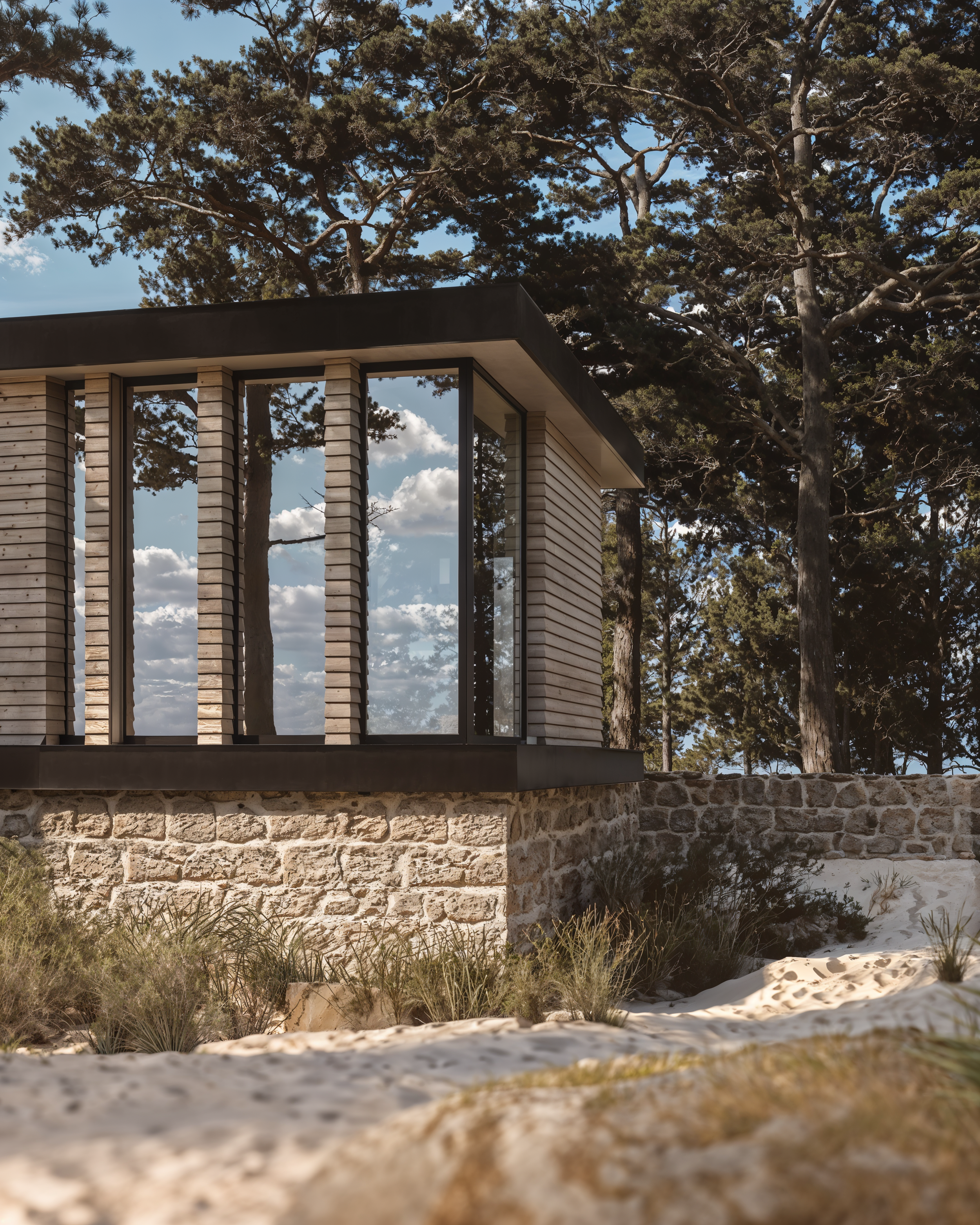 Edifício de arquitetura moderna com estrutura de concreto, rodeado por árvores altas e um ambiente natural com areia e plantas rasteiras em primeiro plano.
