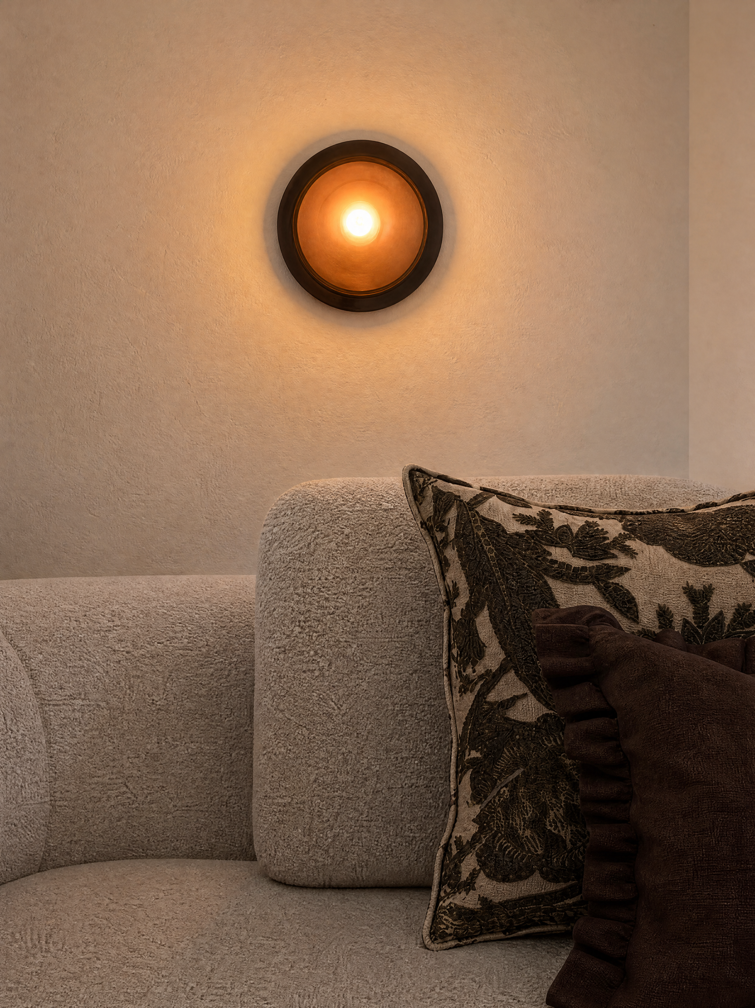 Sofá bege com aconchegante almofada decorativa, ao lado de uma parede de textura suave, com uma luminária de parede circular emitindo luz quente.