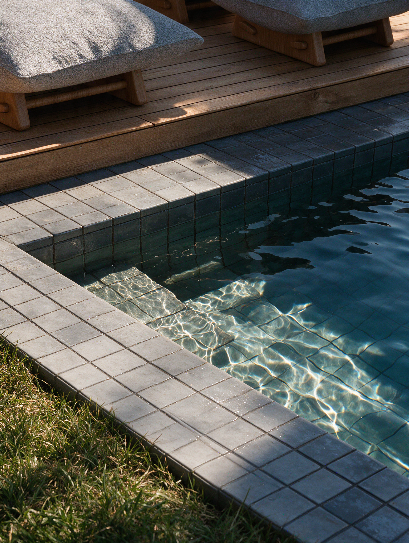Cadeiras com almofadas ao lado de uma piscina com borda de pedra e jardim com grama bem cuidado.
