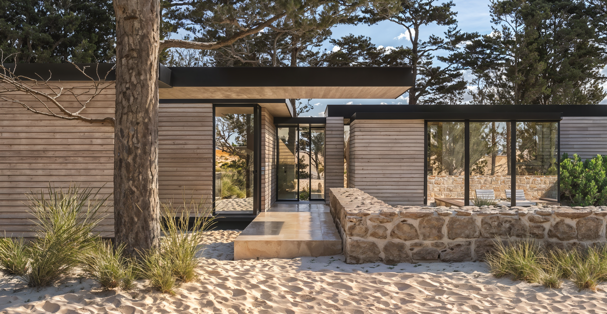 Casa moderna de vidro e madeira, situada na praia, com árvores ao redor, areia e pedras na frente.